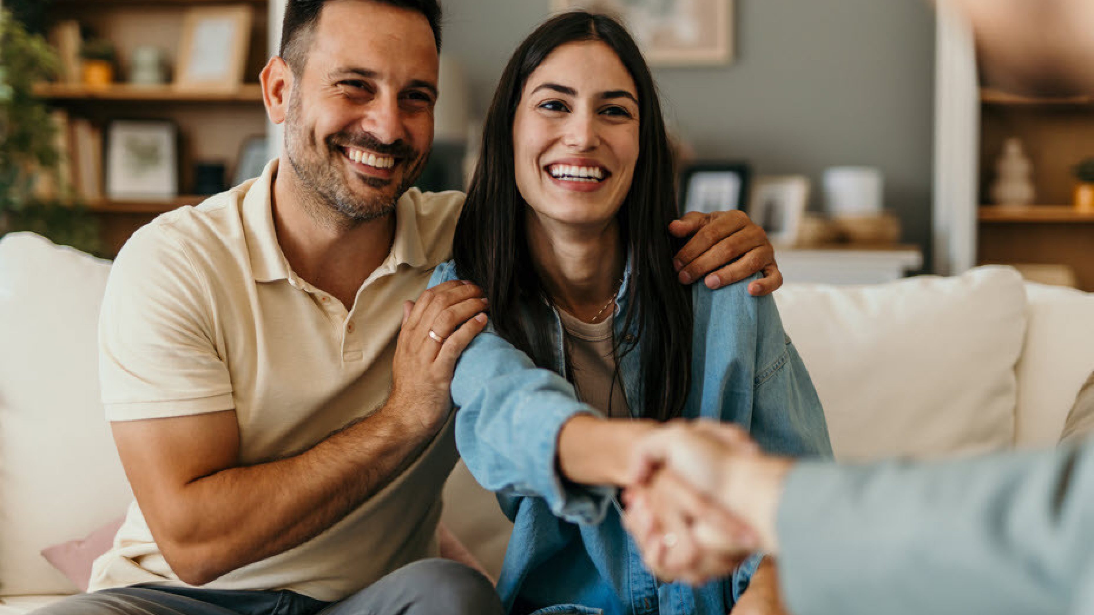 Un homme et une femme sont assis sur le canapé. Tous deux rient. L’homme serre la femme dans ses bras tandis qu’elle serre joyeusement la main de quelqu’un.