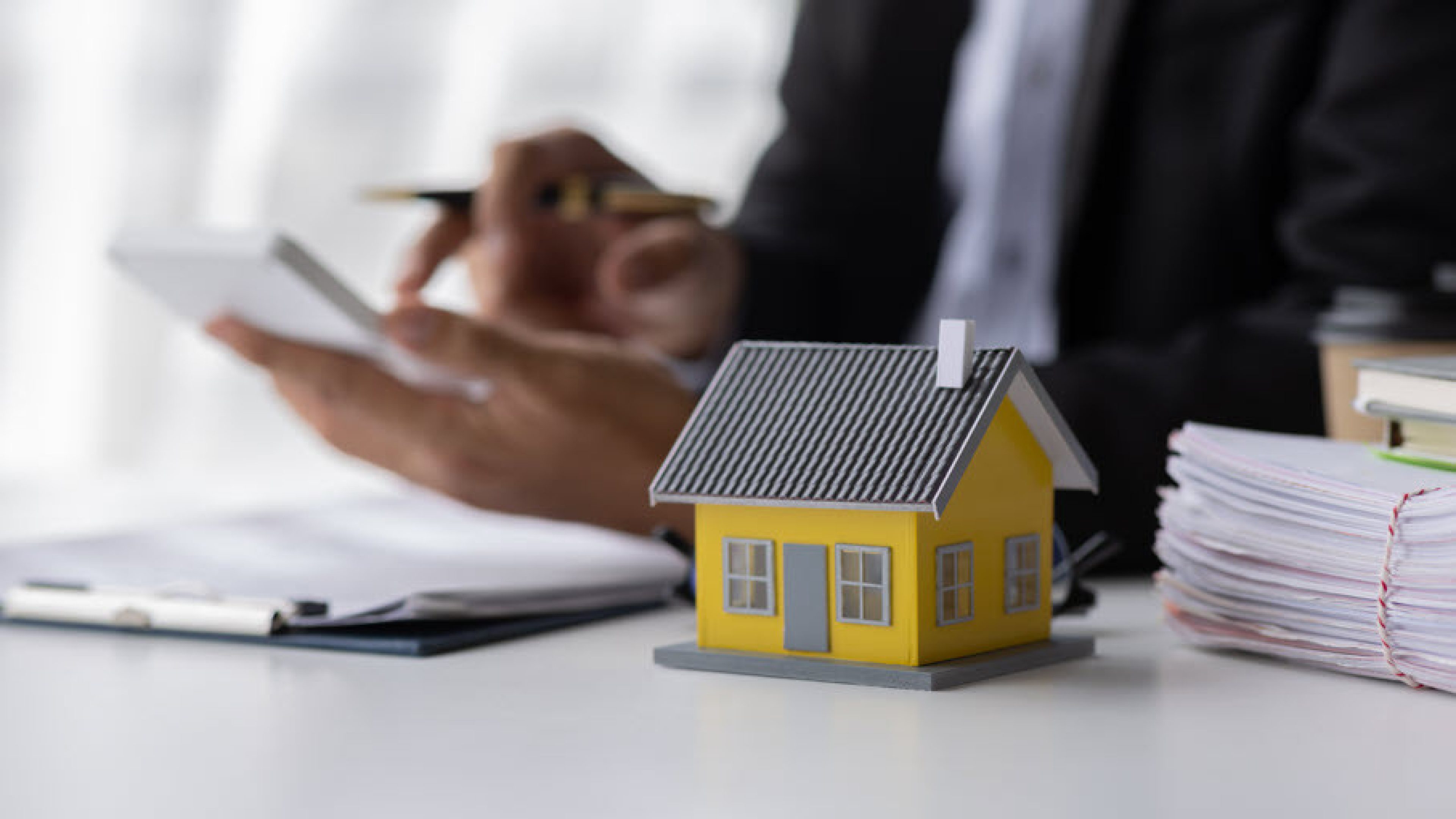 Un conseiller en immobilier est assis à une table et calcule quelque chose sur sa calculatrice. Sur la table se trouvent des documents et une maison modèle jaune.