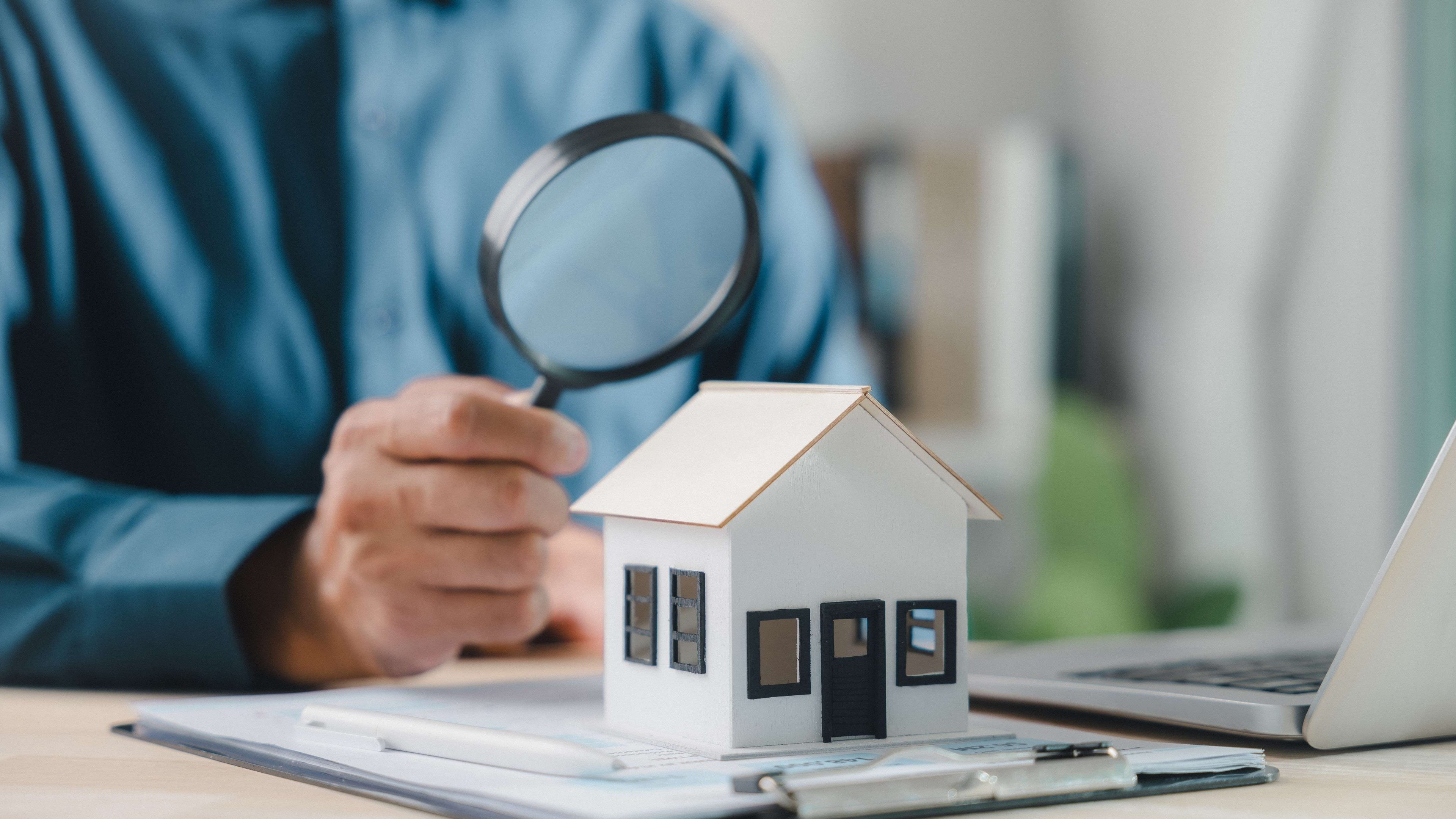 Un homme est assis à un bureau et regarde avec une loupe une maison modèle blanche. Un ordinateur portable et des notes se trouvent également sur la table.
