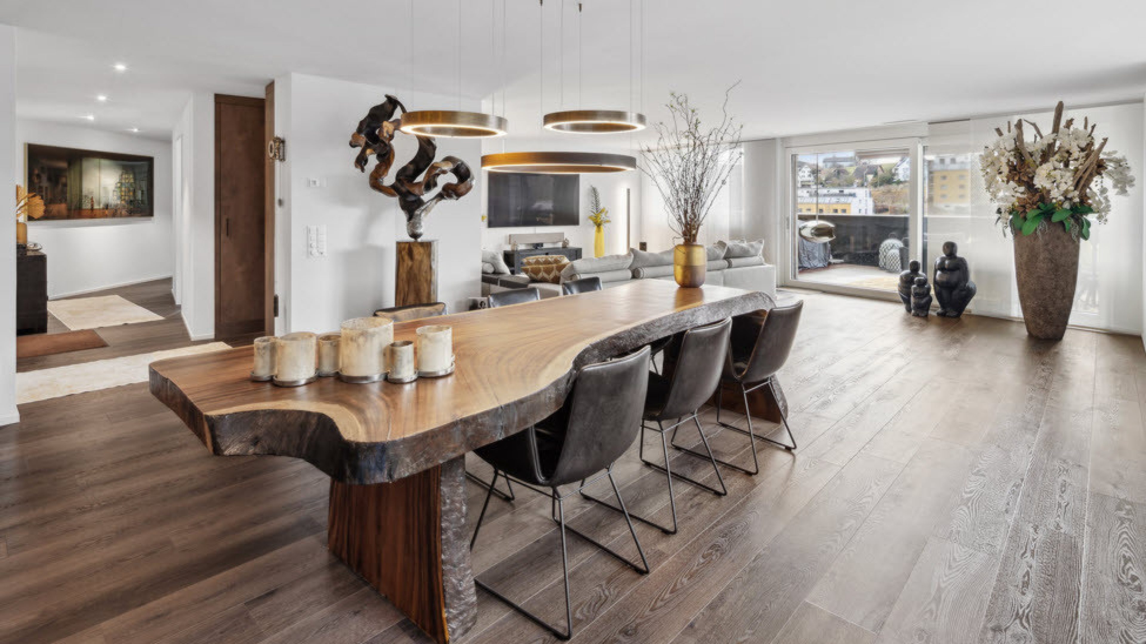 Salon lumineux et moderne. Au centre se trouve une grande table en bois. Le sol est également en bois. Différents vases, grands et petits, avec des fleurs, ainsi que des objets d’art se trouvent dans la pièce.