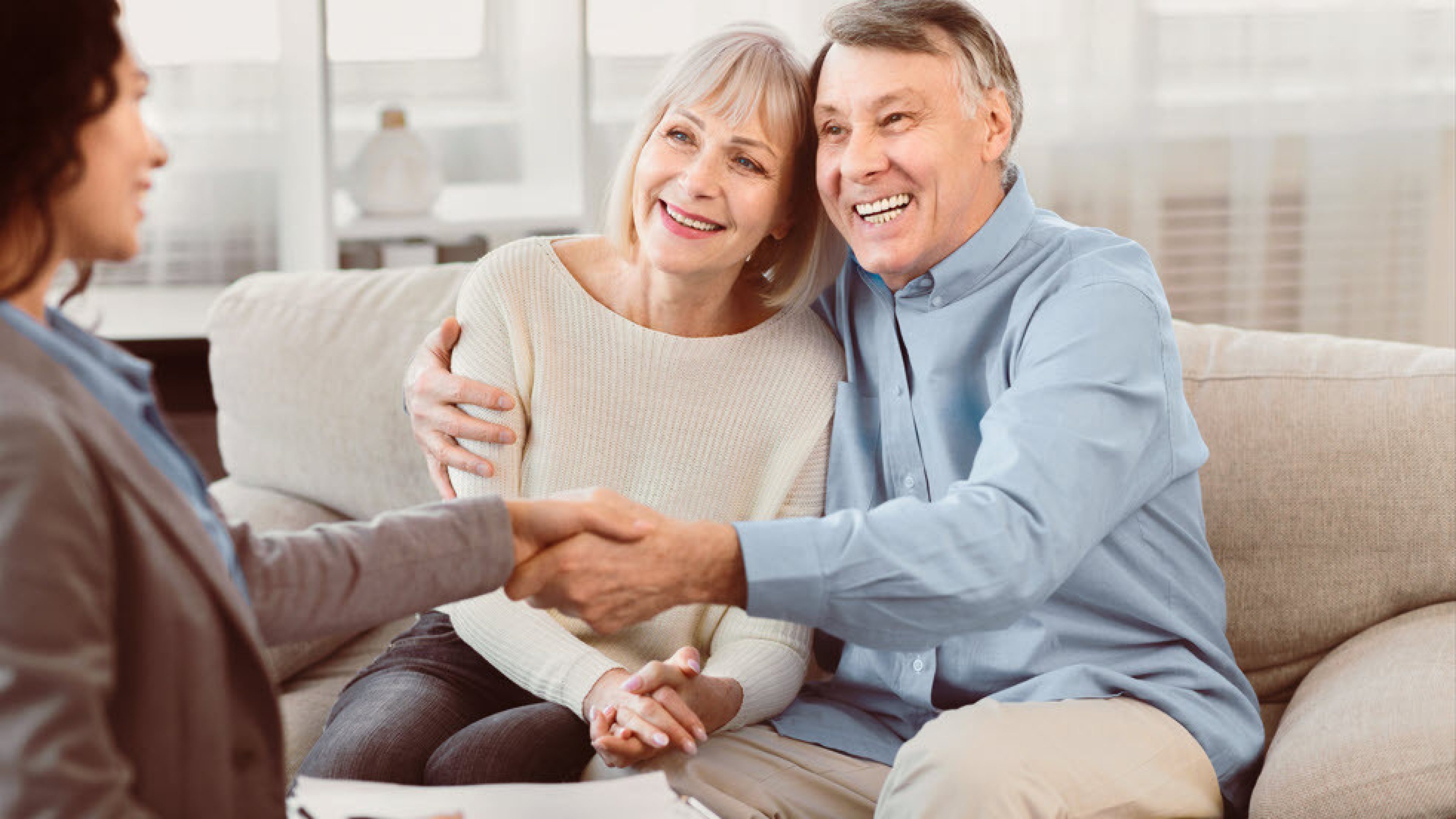 Un couple d’un certain âge est assis sur le canapé. Tous deux sont joyeux et l’homme serre gentiment la main de la conseillère en immobilier.
