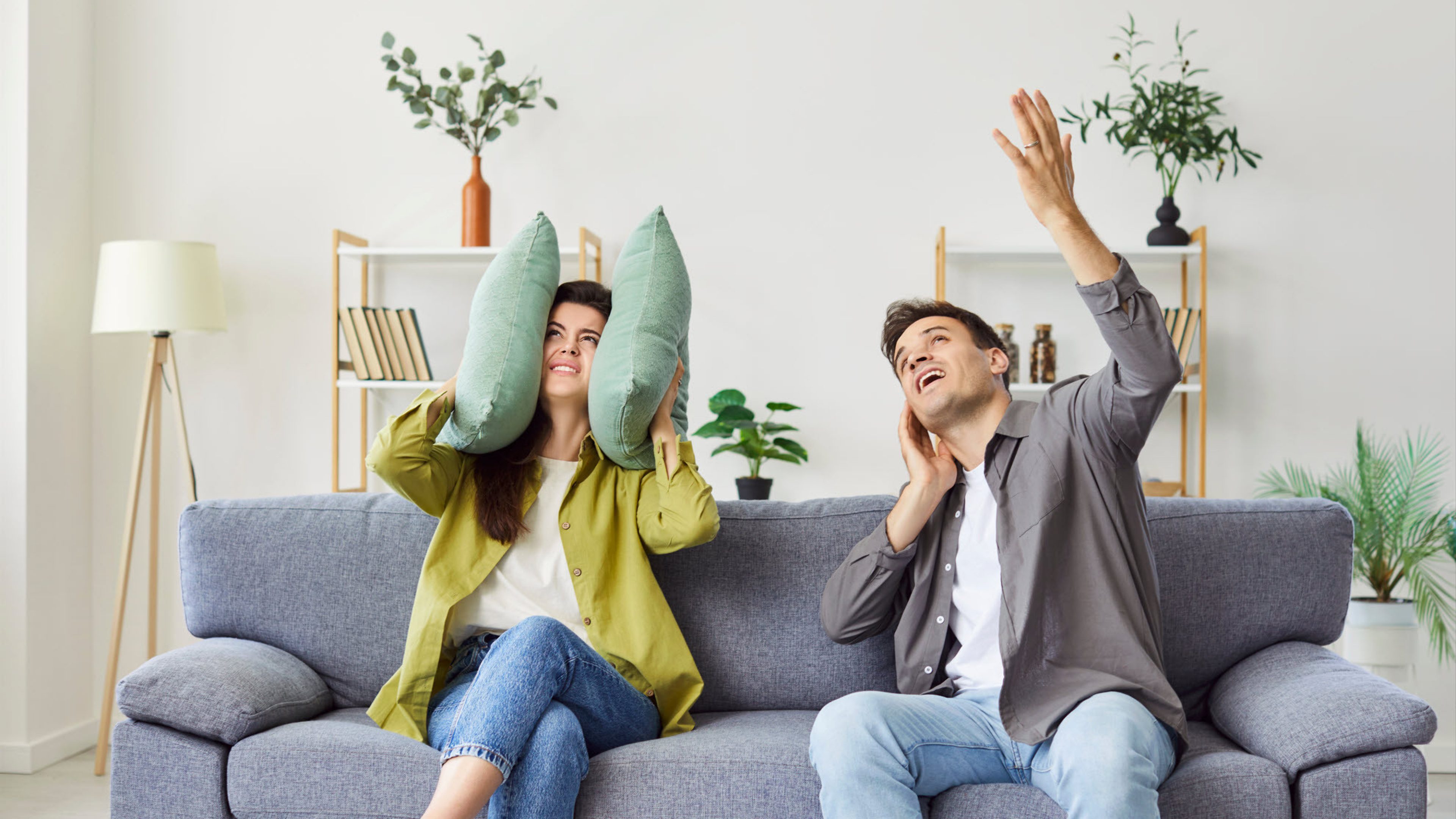 Une jeune femme et un jeune homme sont assis sur leur canapé bleu gris dans le salon lumineux. Tandis que la femme, avec deux coussins vert clair, se tient les oreilles et regarde vers le plafond, l’homme tient également une oreille avec sa main couverte et tend l’autre main vers le plafond et semble se plaindre.