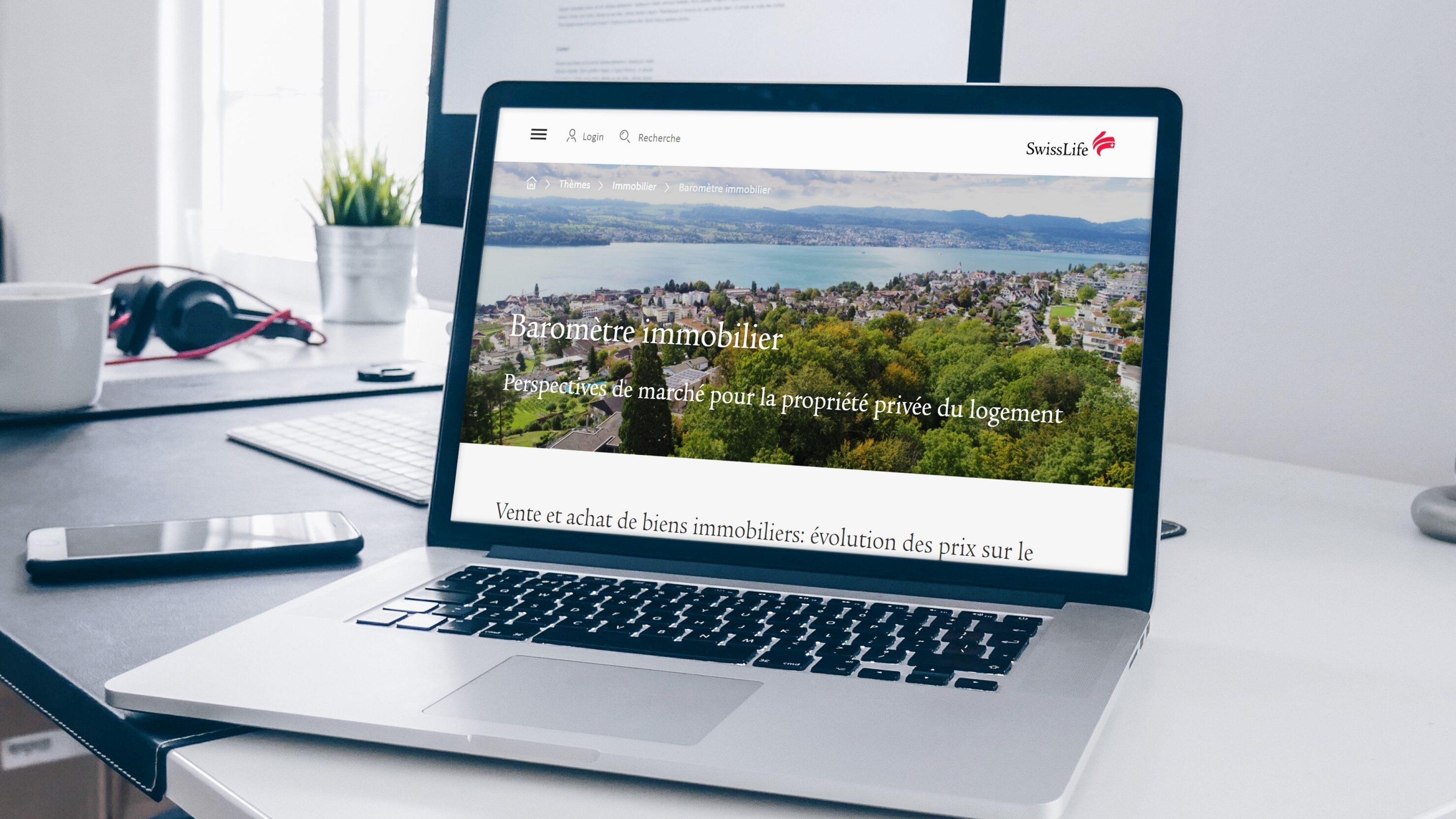 Sur le bureau se trouve un ordinateur portable ouvert sur lequel figure la page du baromètre immobilier Swiss Life: un lac, des maisons et des arbres.