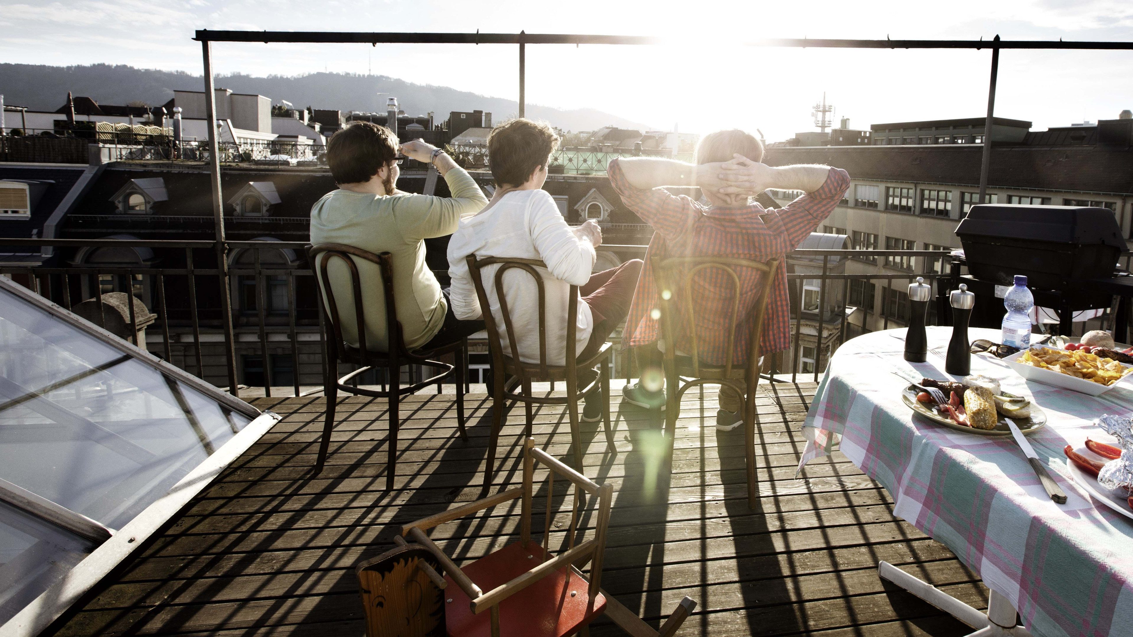 Trois personnes sont assises sur une terrasse ensoleillée et profitent du soleil du soir. A côté se trouve une table couverte. Au premier plan se trouve un cheval à bascule.