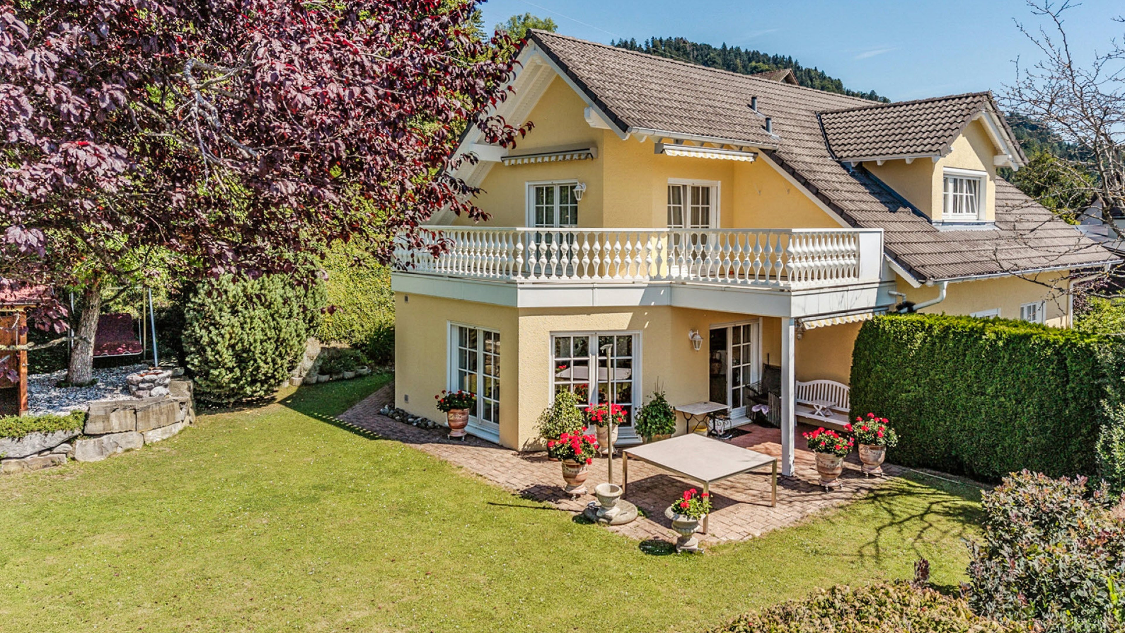 Maison individuelle jaune accueillante avec balustrade blanche et fenêtres blanches. Sur la terrasse se trouvent une table et des pots de fleurs rouges. La maison est entourée d’un espace vert soigné et d’arbres.