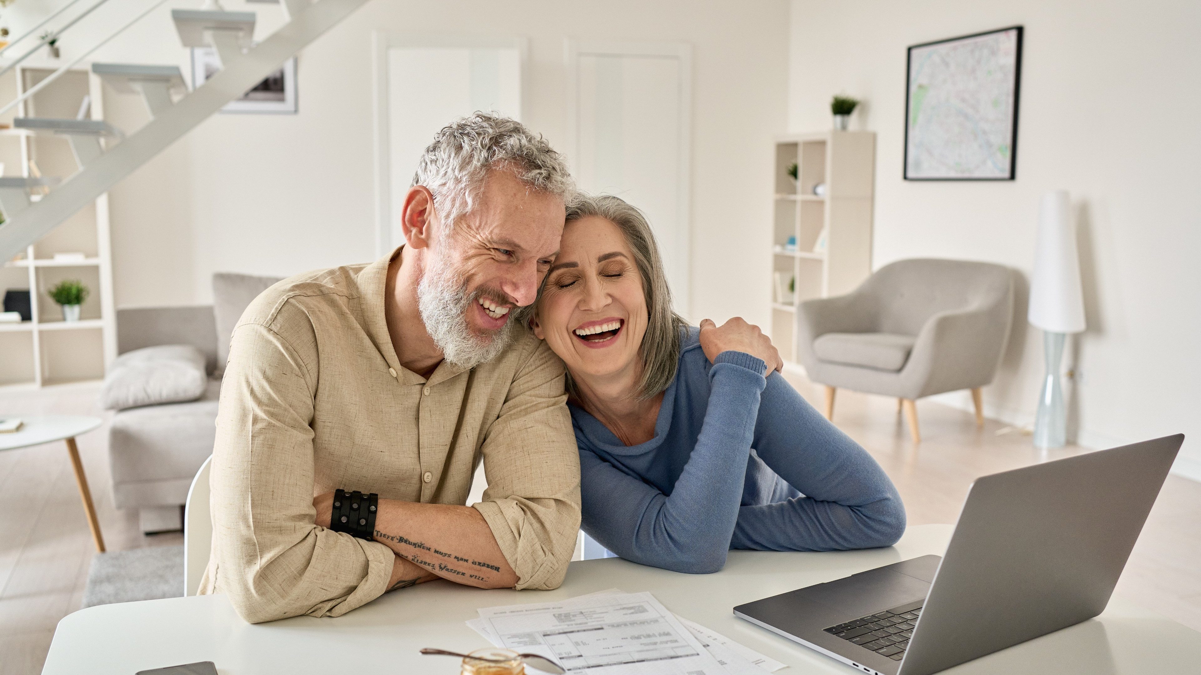 Happy mature older family couple laughing, bonding sitting at home table with laptop. Smiling middle aged senior 50s husband and wife having fun satisfied with buying insurance, paying bills online.