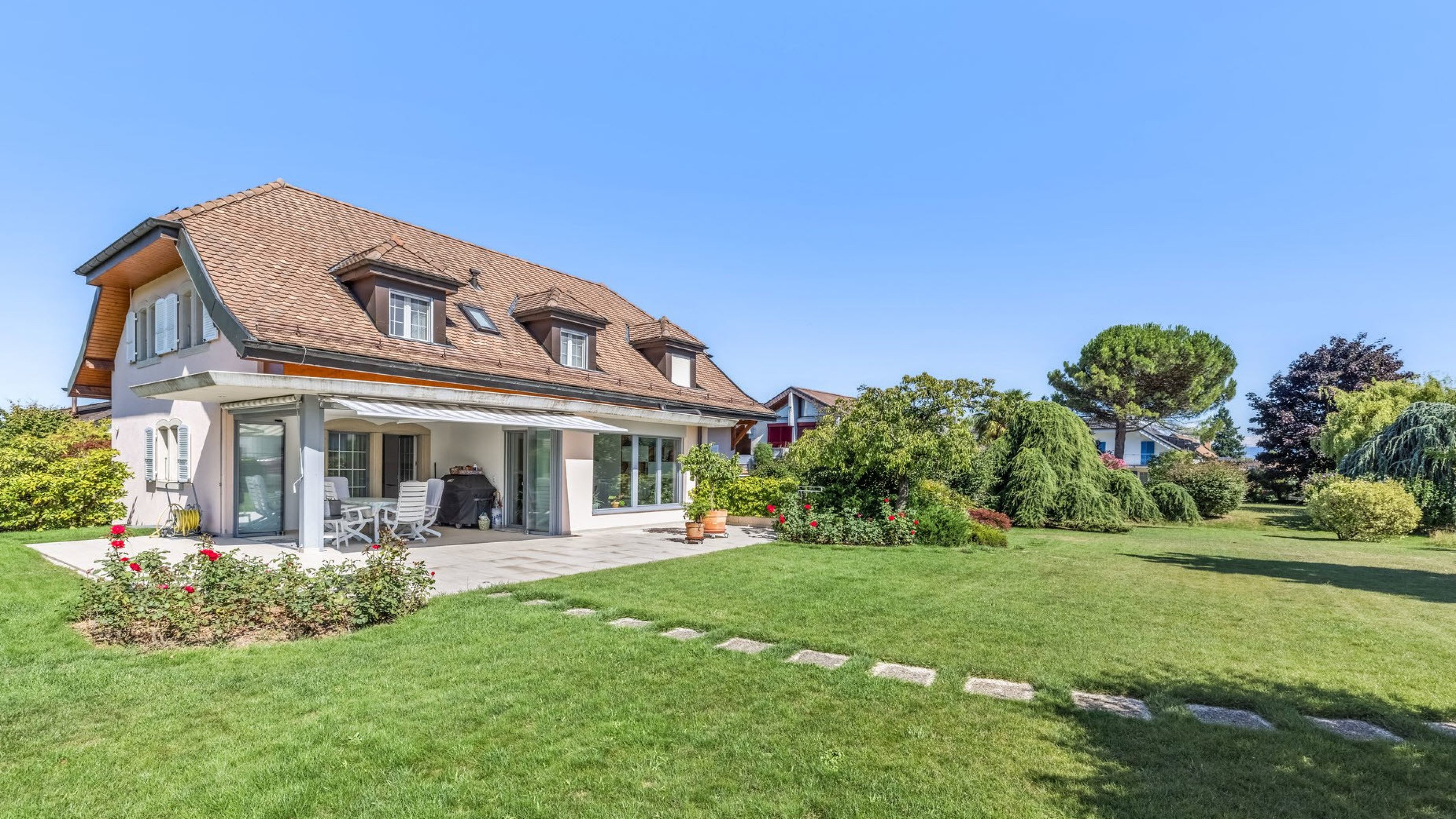 Maison individuelle lumineuse et conviviale avec une grande pelouse fraîchement tondue. Dans le jardin, il y a des roses rouges et divers arbustes et arbres. Le ciel est bleu, le soleil brille.