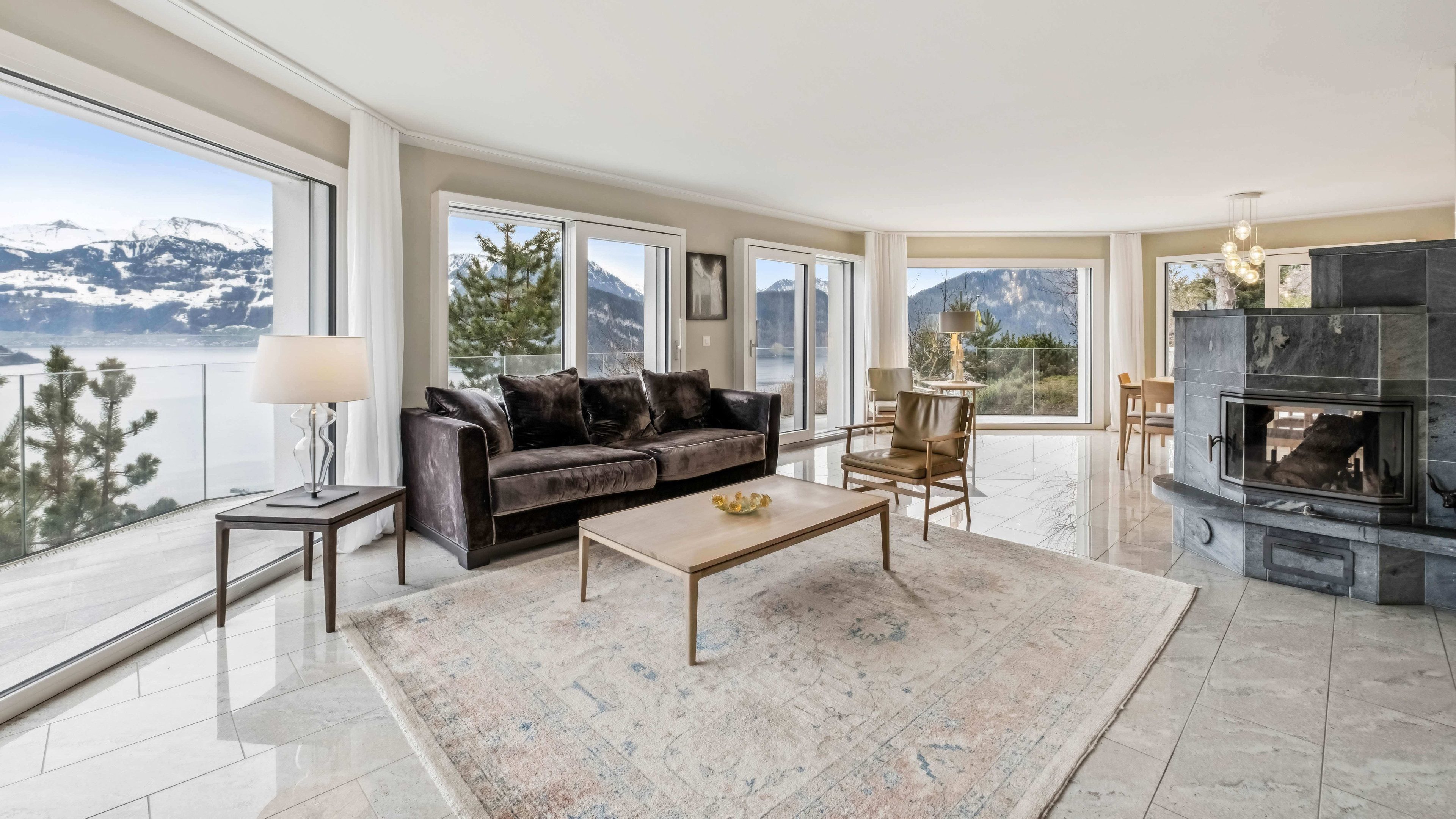 Salon lumineux avec carrelage et grandes baies vitrées. Le salon dispose d’un canapé ainsi que de diverses tables et chaises. A travers les fenêtres, on voit des montagnes enneigées.