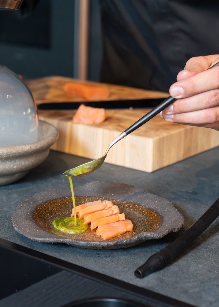 Quelqu’un prépare un plat dans une cuisine moderne. La personne verse une sauce verte au moyen d’une cuillère sur des tranches de saumon disposées sur une assiette. En arrière-plan: une planche à découper en bois sur laquelle est posé du saumon et une cloche de verre remplie de fumée.