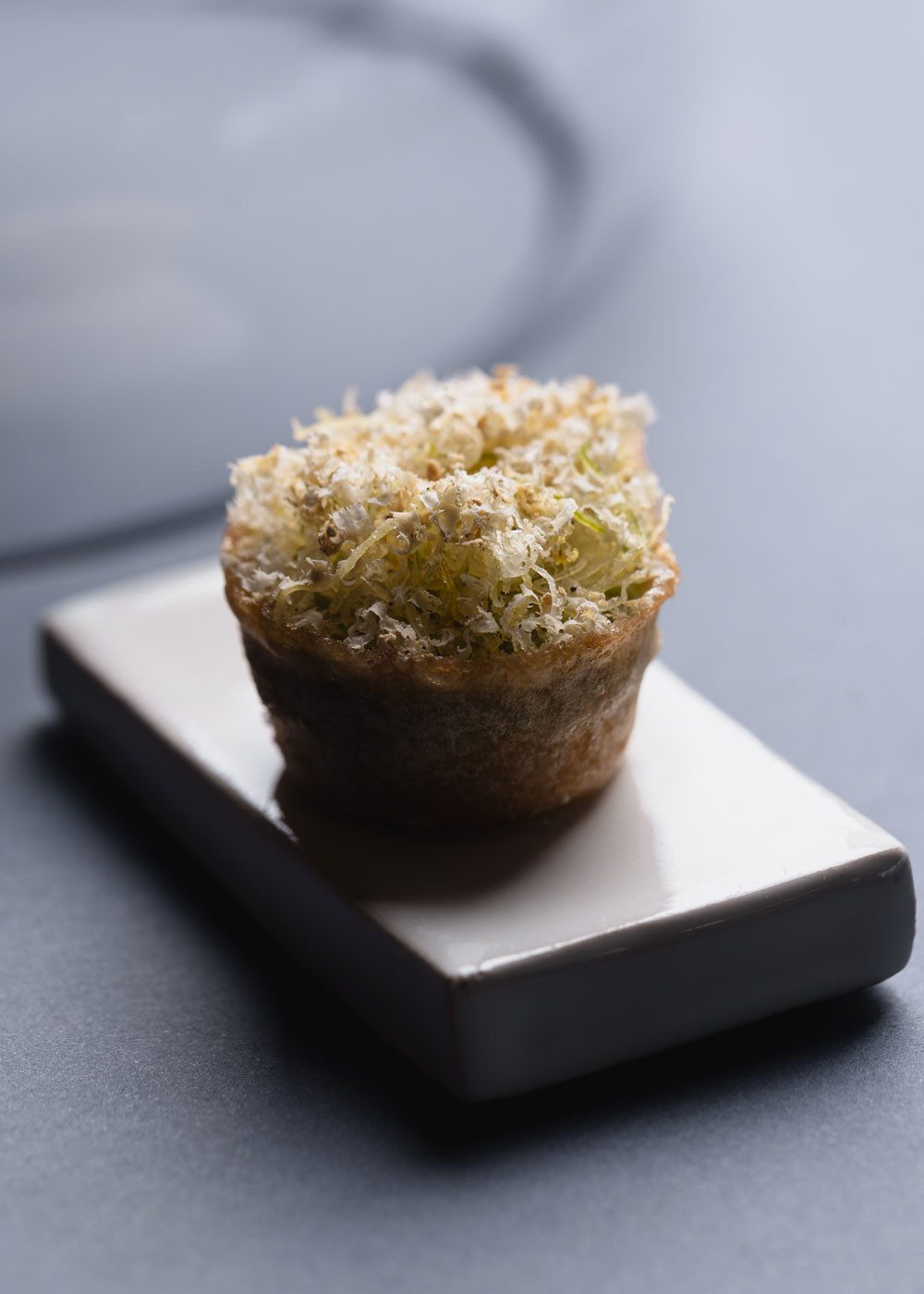 Petit plat savamment décoré sur une assiette en céramique carrée. Le plat ressemble à un fond de tartelettes croustillant garni d’un ingrédient râpé. Il se tient devant un fond gris qui souligne le design élégant des plats.