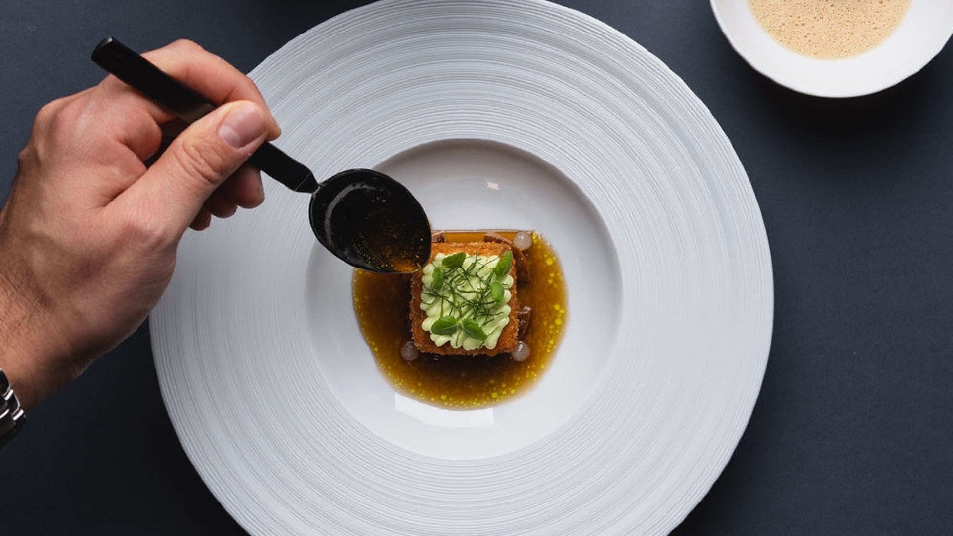 Gros plan d’une assiette avec un plat savamment décoré. Une main verse une sauce sur l’assiette à l’aide d’une cuillère. A côté: un autre petit bol contenant un autre ingrédient.