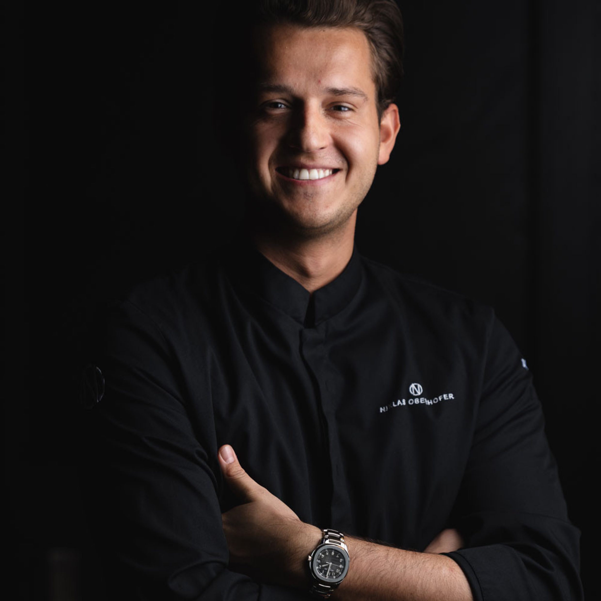 Portrait d’un jeune homme aux cheveux bruns courts affichant un sourire amical sur fond noir. Il porte une veste de cuisinier noire avec son nom brodé dessus sur la poitrine et une montre. Ses bras sont croisés.