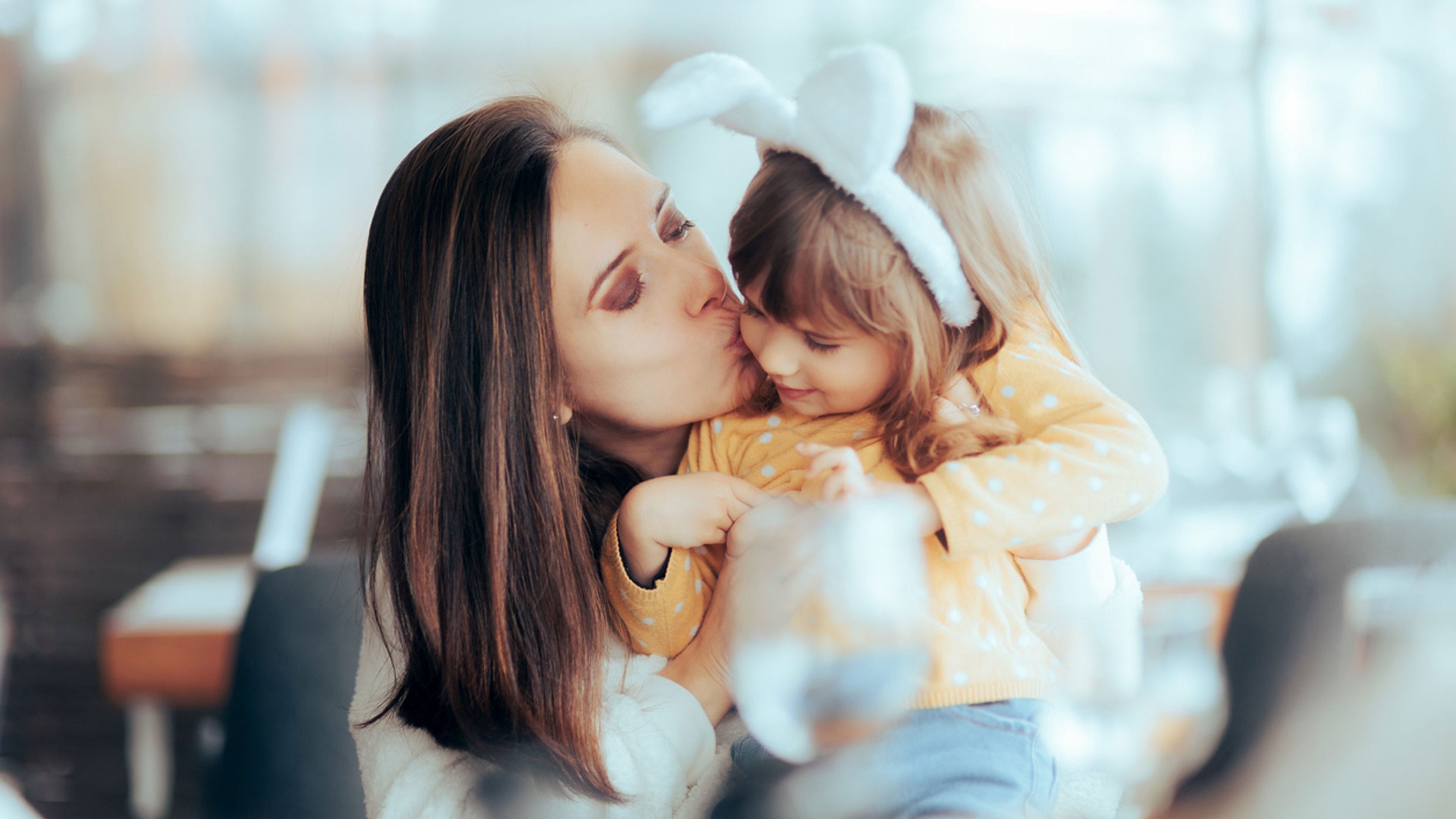 Une mère aux cheveux bruns embrasse sa petite fille portant des oreilles de lapin blanches.