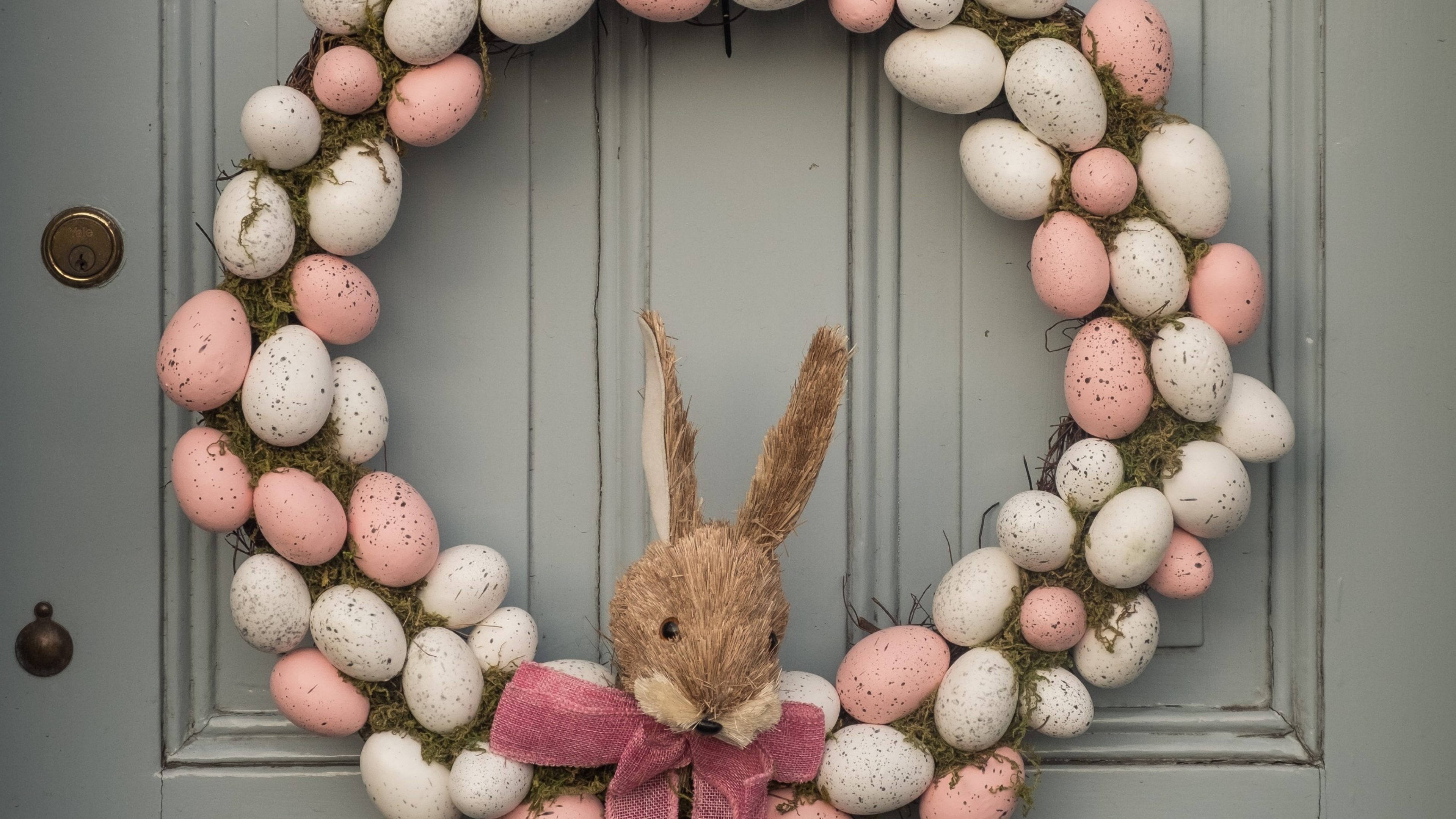 Une couronne de porte composée d’œufs de Pâques de couleurs pastel et d’un lapin décoratif accrochée à une porte en bois grise.