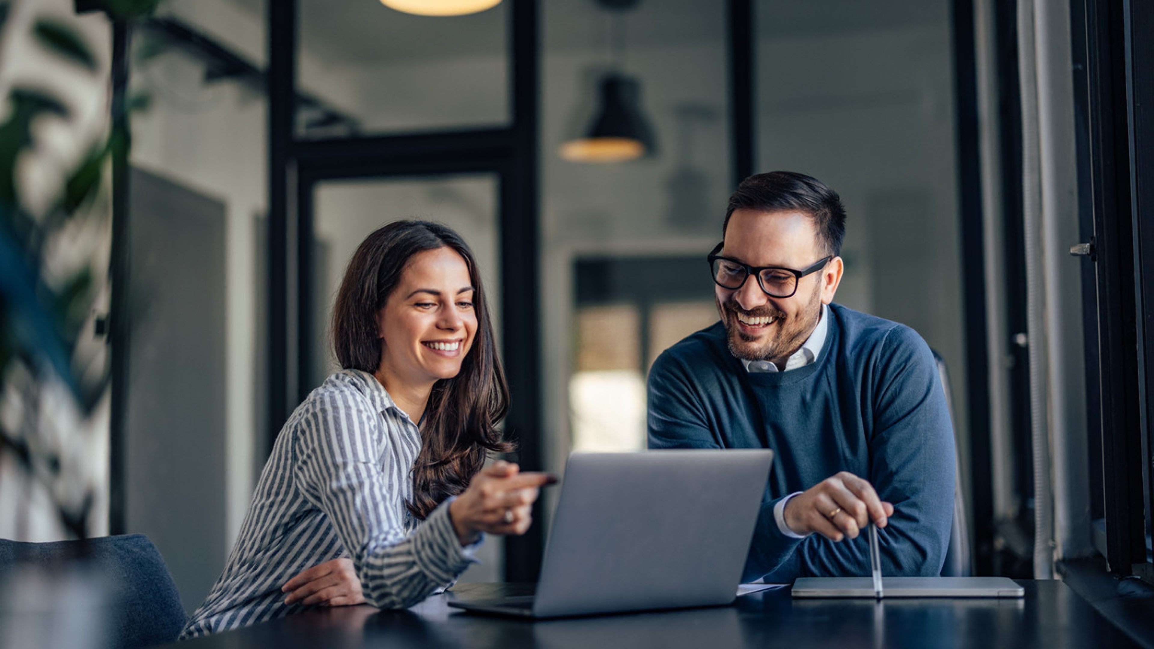 Un homme et une femme sont assis à une table dans un bureau moderne et regardent ensemble un ordinateur portable. Ils rient et ont l’air détendus. La femme porte un chemisier à rayures et l’homme, un pull bleu. En arrière-plan, de grandes fenêtres et des lampes de bureau sont visibles. La pièce est bien éclairée et décorée dans un style moderne.