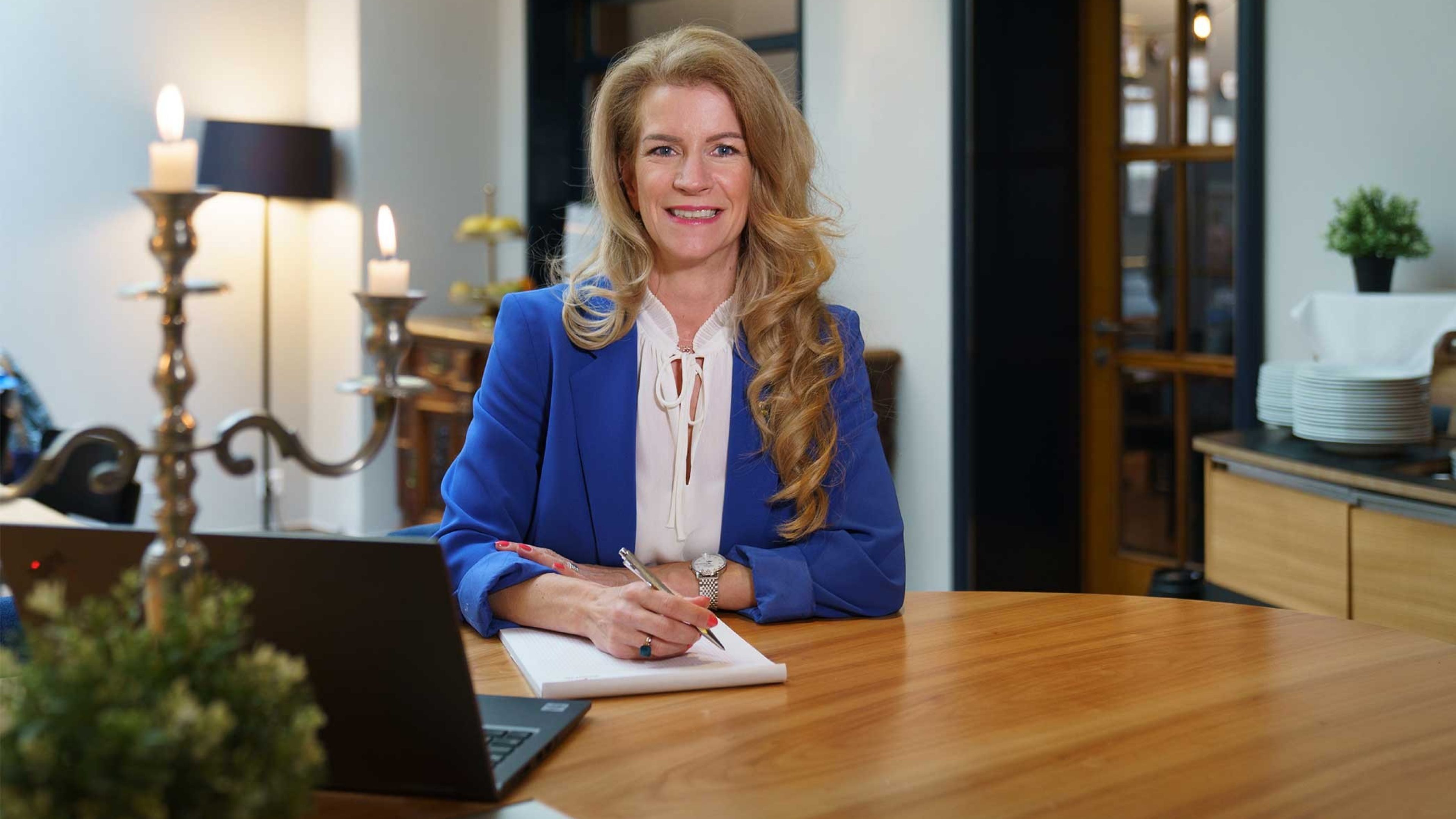 Une femme blonde souriante est assise à un bureau en bois. Elle porte un chemisier blanc et une veste bleu royal. Elle tient un stylo dans sa main droite et a un bloc-notes devant elle. Un chandelier argenté et une plante sont posés sur la table.
