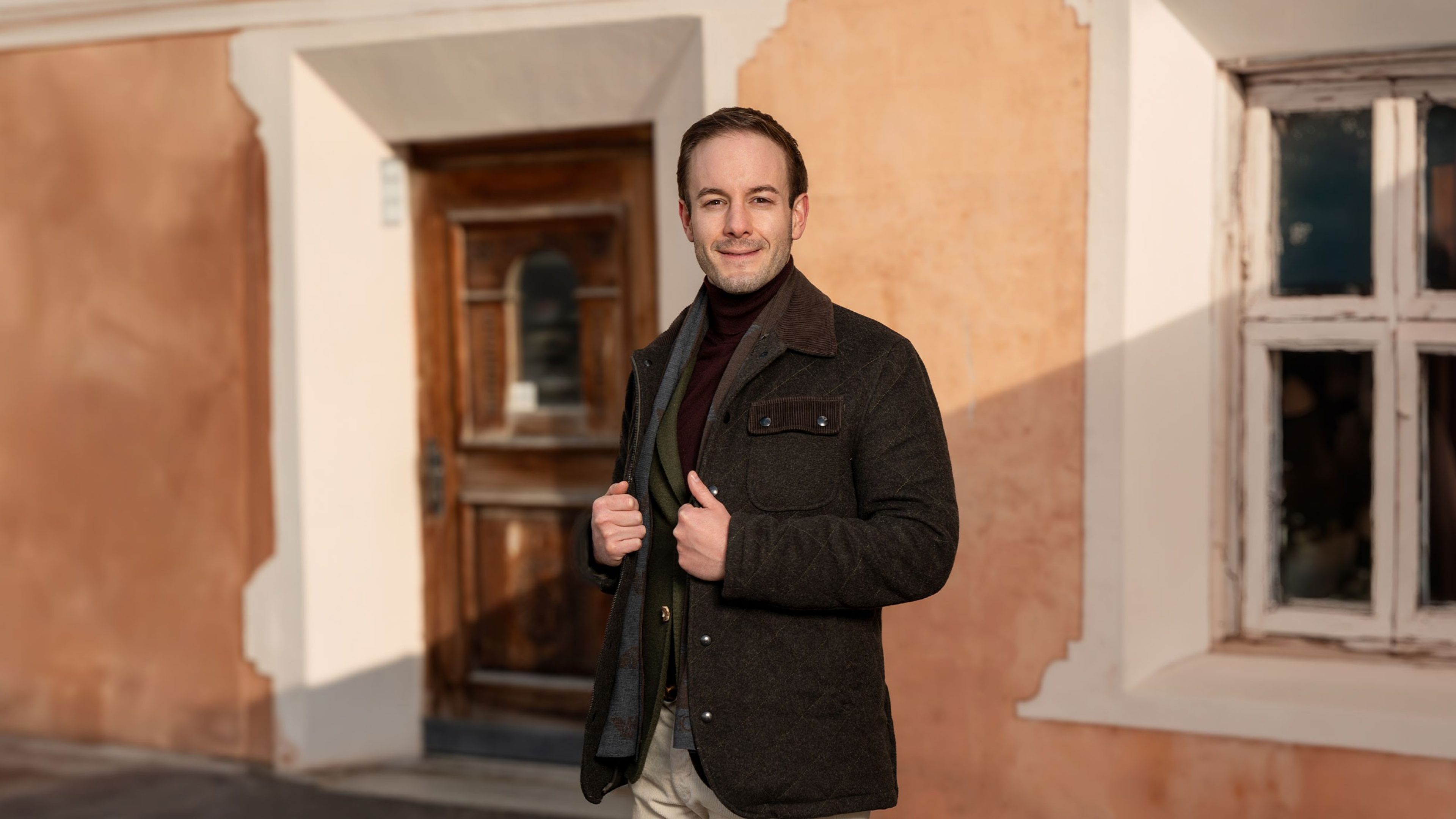 Un homme se tient devant une façade de maison décorée en Engadine. La façade de couleur abricot présente des ornements en stuc blanc autour de la fenêtre à droite et une porte rustique en bois au centre. L’homme a les cheveux courts, une barbe de trois jours et une veste marron foncé.