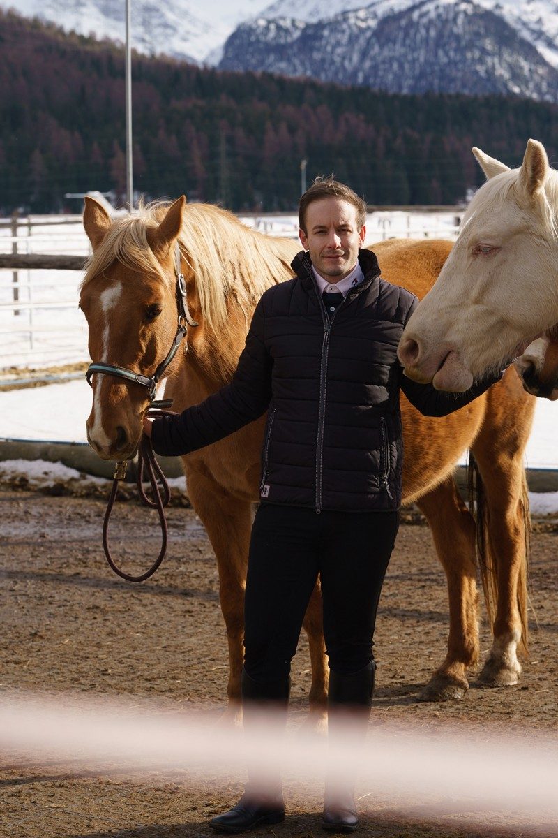 Un homme se tient debout avec deux chevaux, avec des montagnes en arrière-plan. A sa gauche, il tient par les rênes un cheval brun au front blanc et à la crinière claire. La tête d’un cheval blanc est visible à droite. L’homme a les cheveux courts et une barbe de trois jours; il porte une doudoune sombre, un pantalon noir et des bottes d’équitation noires.