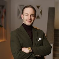 Mann steht in einem Treppenhaus und lächelt in die Kamera. Der Mann hat kurze Haare, einen Dreitagebart und trägt eine dunkelgrüne Jacke.