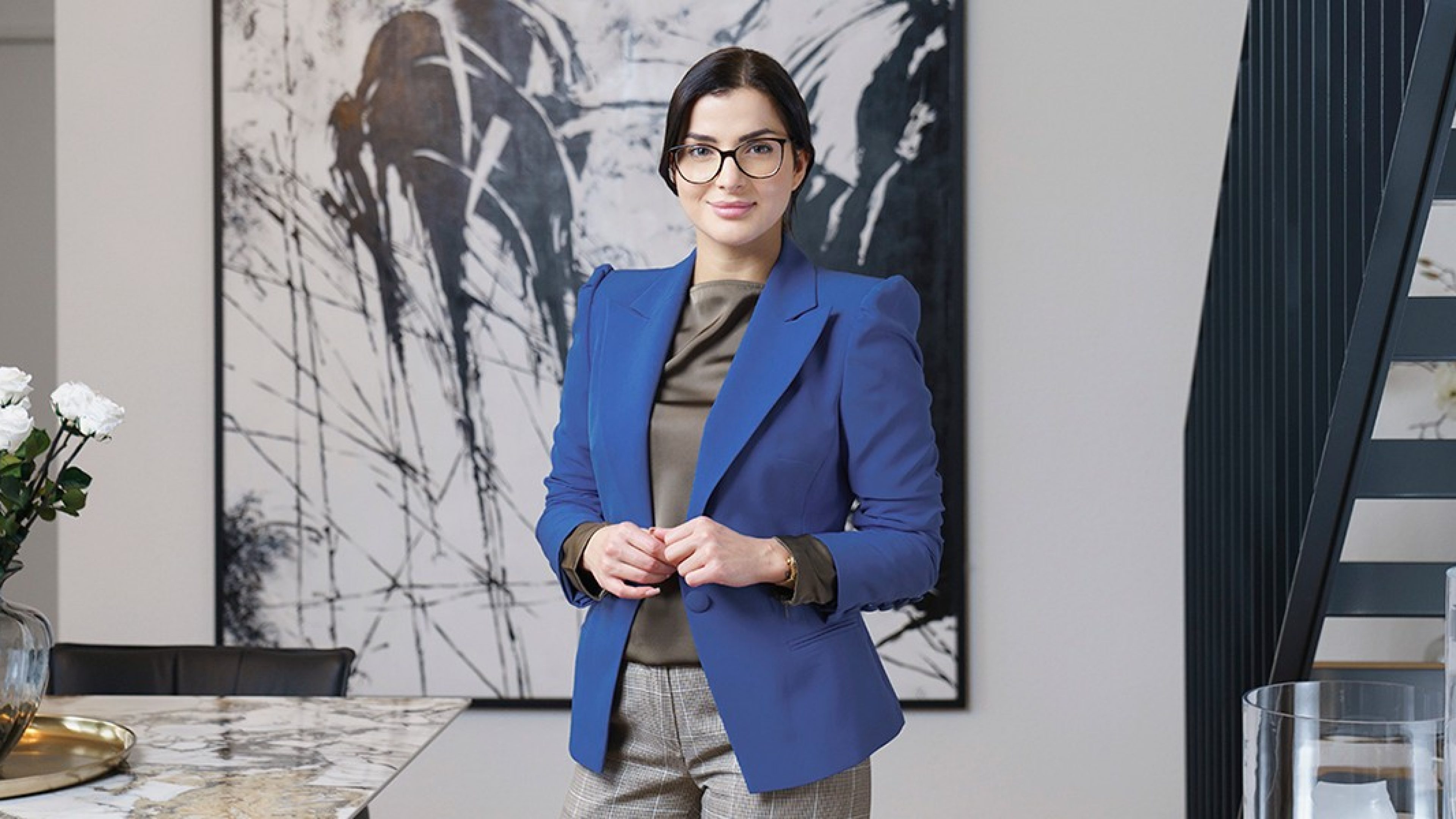 Une femme avec des lunettes en tenue de bureau se tient devant une œuvre d’art et regarde la caméra
