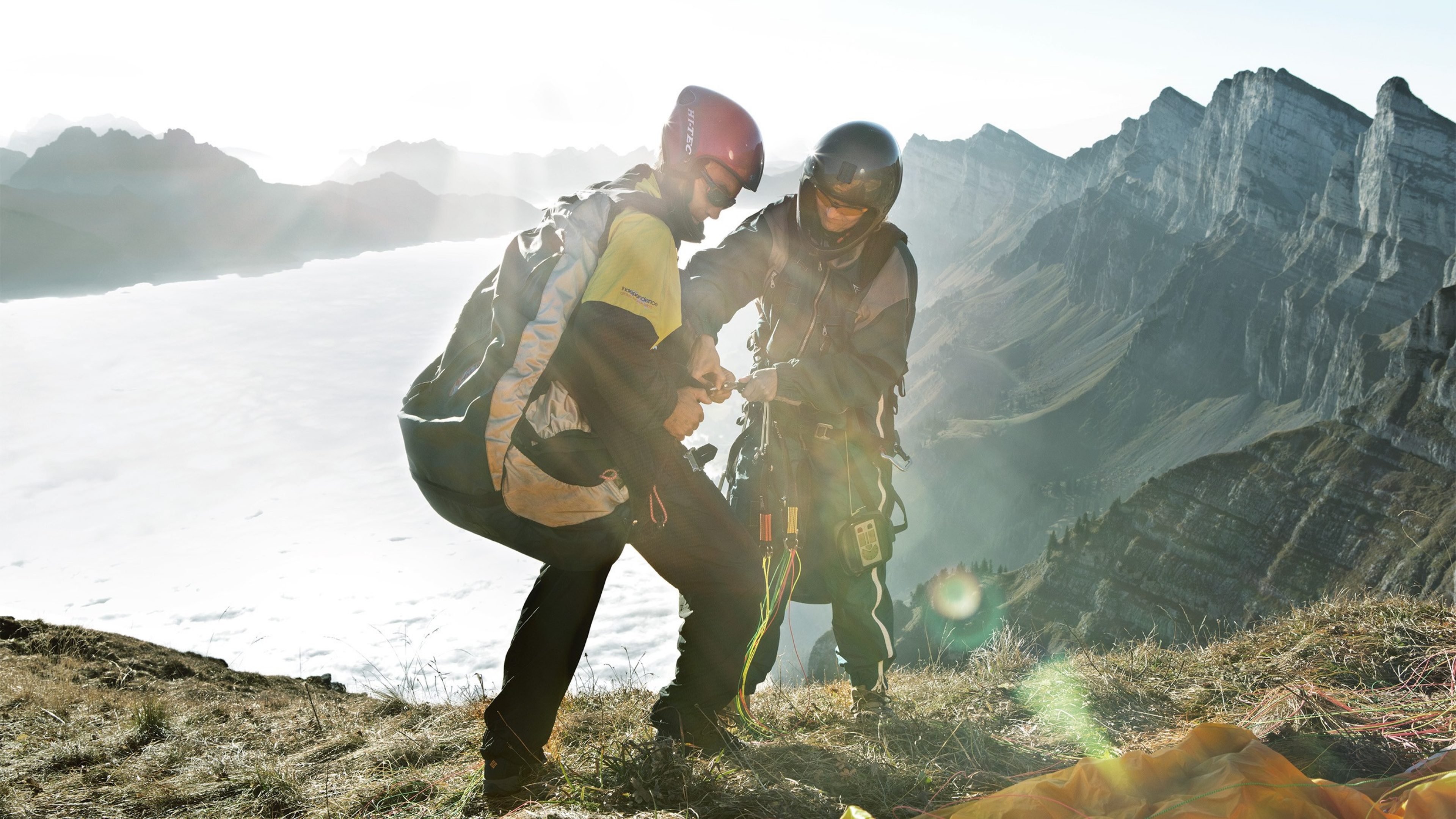 Zwei Personen, bereit für einen Fallschirmsprung, stehen an einem steilen Bergabhang mit Blick auf eine atemberaubende Landschaft. Das Bild vermittelt ein Gefühl von Abenteuer und Adrenalin.