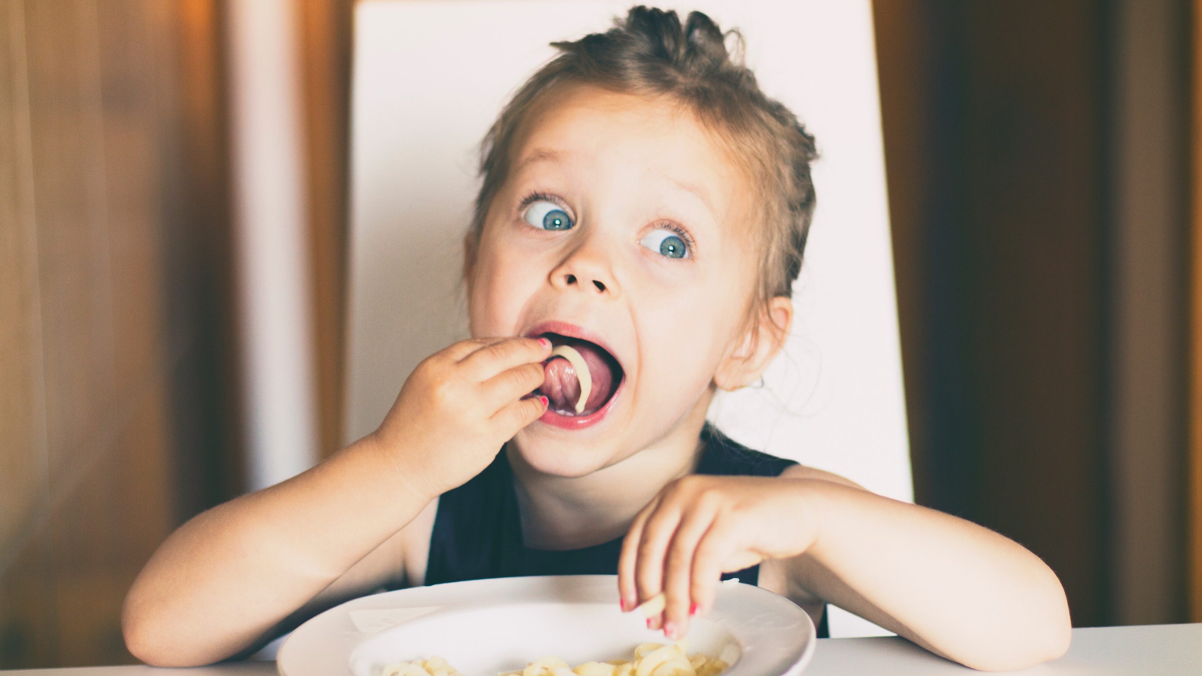 Three years old girl sitting at the table in the kitchen and eating pasta.