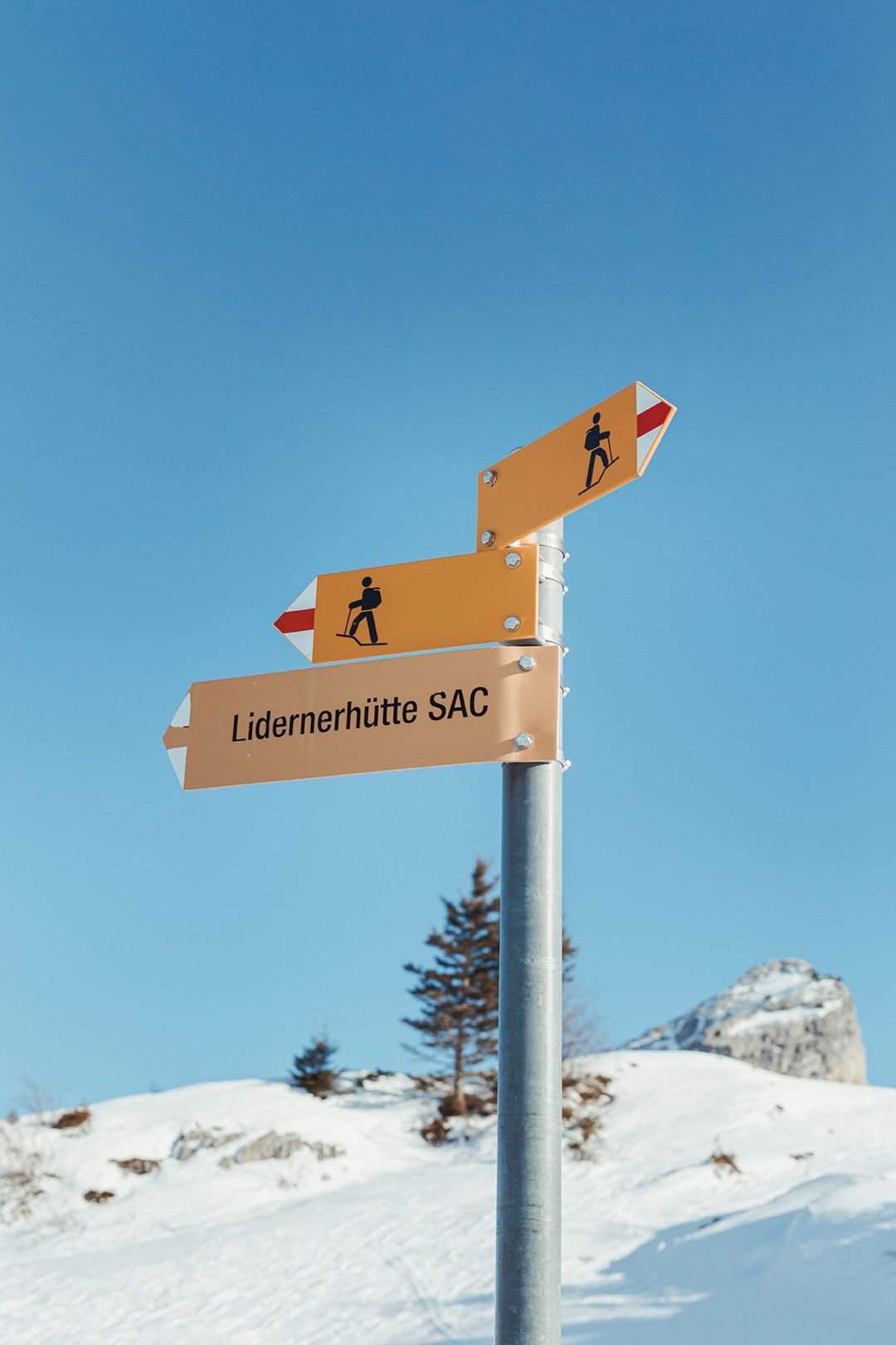 Panneaux de randonnée dont l’un indique la cabane «Lidernenhütte SAC». En arrière-plan, on voit un ciel bleu et un paysage alpin enneigé.