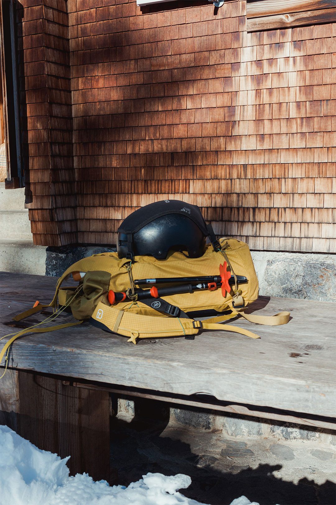 Un sac à dos de randonnée et un casque de snowboard sont posés sur la terrasse d’un chalet de montagne.