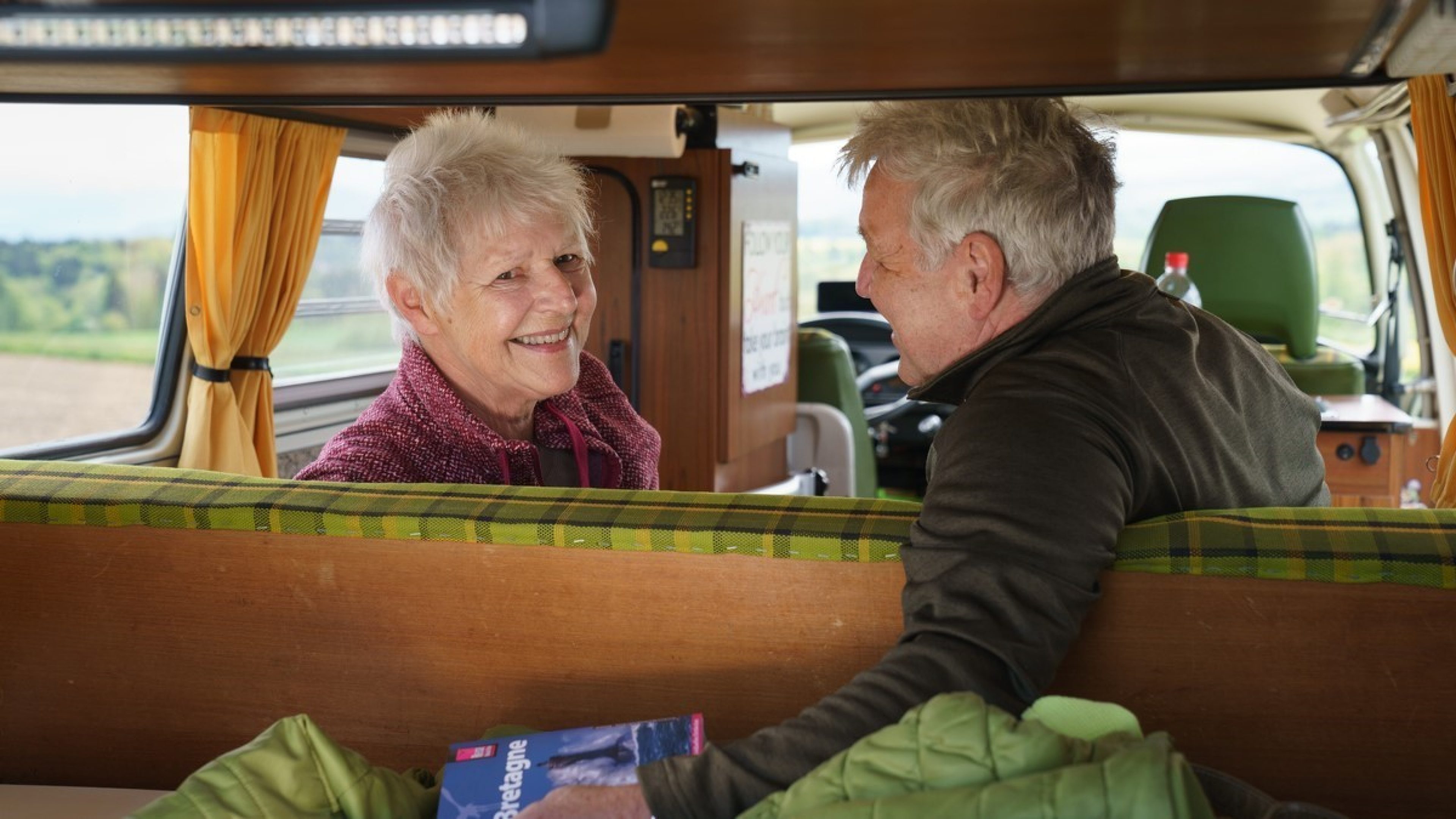 Kurt et Monica sont assis sur le canapé à l’arrière de leur bus. On les voit depuis le coffre. Monica sourit à l’objectif, tandis que Kurt la regarde et tient un livre de voyage sur l’Angleterre dans la main.