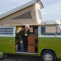 Le bras appuyé sur un cadre en bois, Kurt rit et regarde à travers la porte latérale ouverte de son bus VW couleur vert mousse. Le toit du bus VW est surélevé en diagonale.