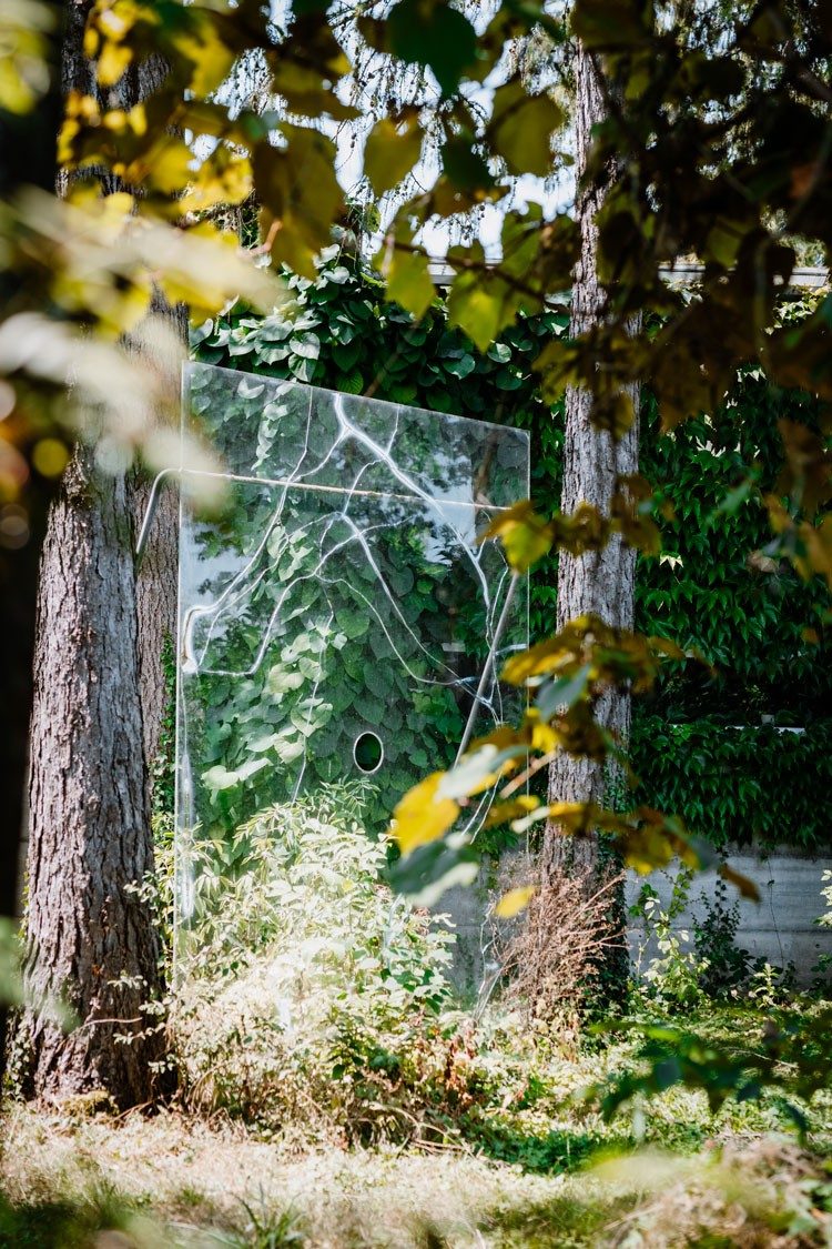 Une œuvre d’art en verre est installée entre deux arbres du jardin. Sa paroi en verre est parcourue de rayures incurvées et comporte un trou en son centre.