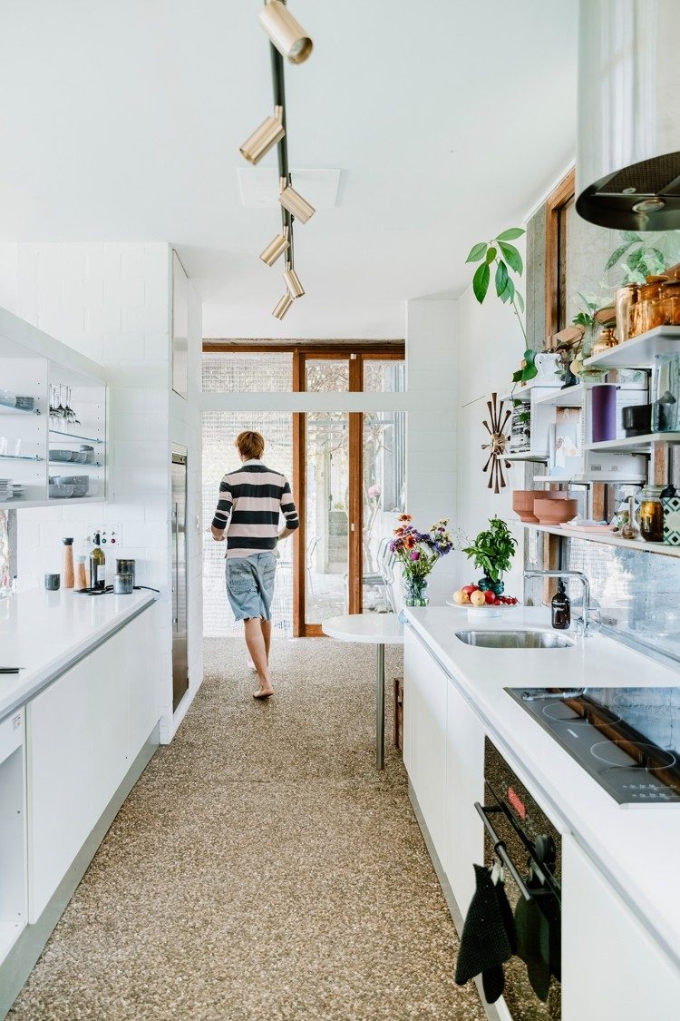 Dans une cuisine lumineuse ornée de plantes et équipée de divers ustensiles, un jeune homme vêtu de bleu et blanc marche vers une porte-fenêtre coulissante qui mène au jardin. Un long plafonnier à spots décore le plafond.