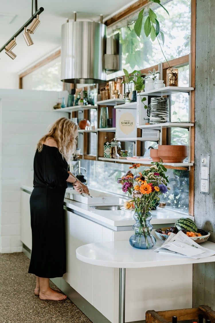 Une femme aux cheveux blonds ondulés vêtue d’une robe noire prépare quelque chose dans sa cuisine. La cuisine est décorée de divers récipients et d’un vase de fleurs colorées.
