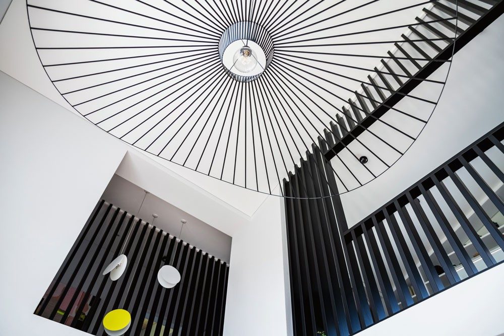 Une grande lampe de plafond aux éléments noirs en filigrane s’intègre parfaitement dans une cage d’escalier avec rampe et garde-corps. 