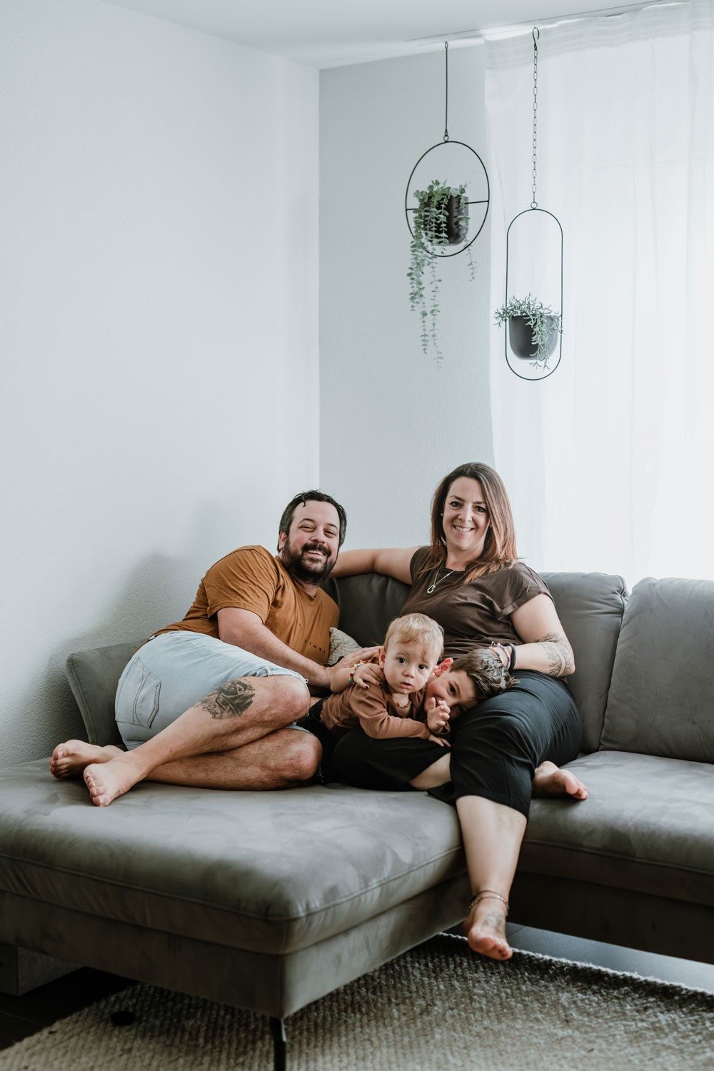 Une femme, un homme et deux jeunes enfants sont allongés sur un canapé gris. Au-dessus d’eux, des plantes sont suspendues. Les deux adultes sourient à la caméra.