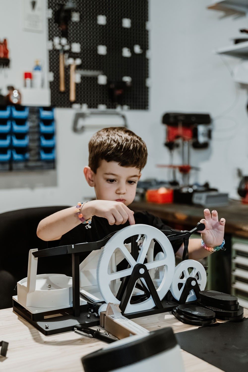 Un petit garçon joue avec un jouet noir et blanc composé de roues et de circuits. Le garçon est concentré. Il a les cheveux bruns, porte un t-shirt noir et deux bracelets colorés.
