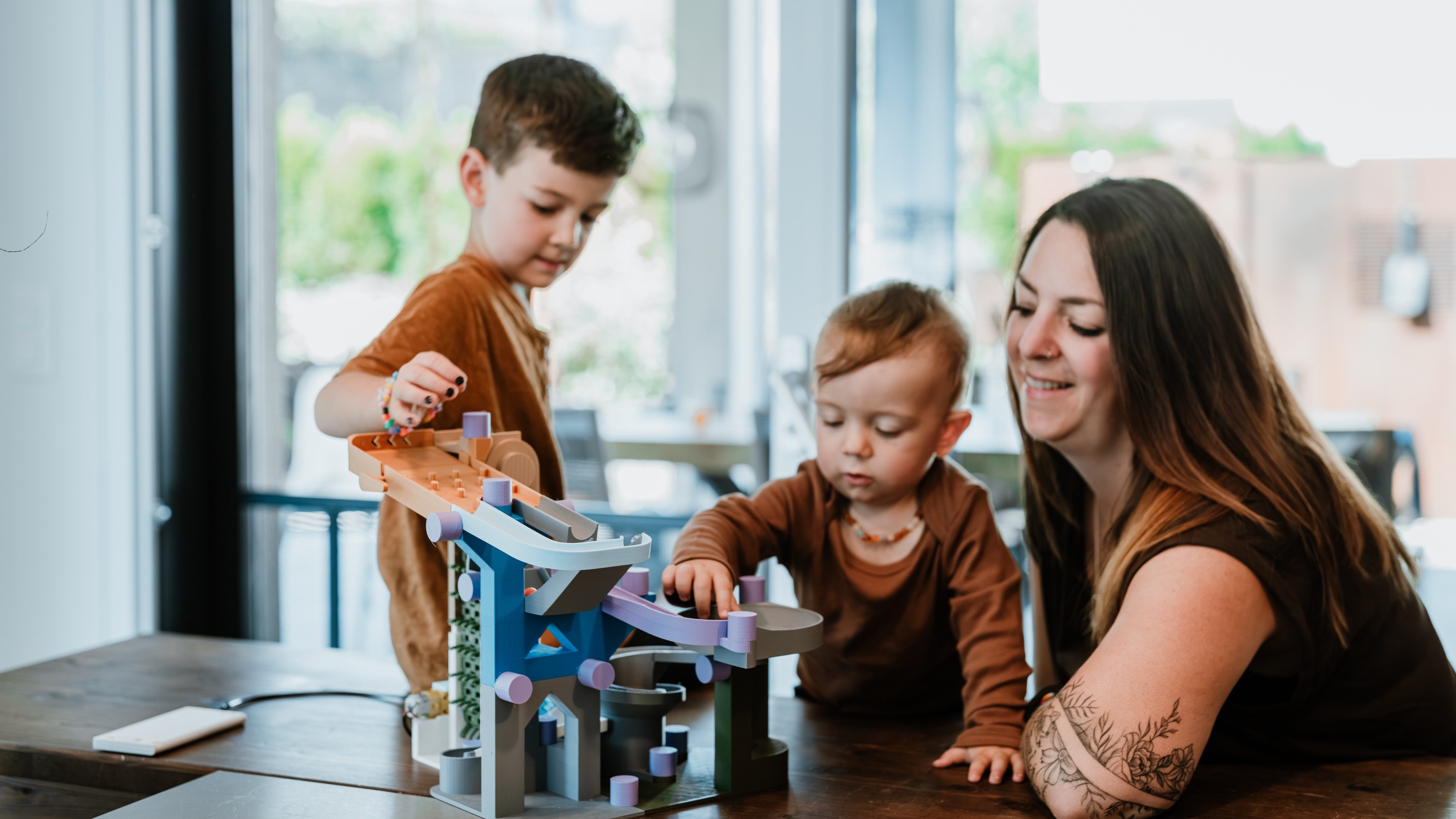 Une femme à l’avant-bras tatoué observe en souriant deux enfants jouant à une table avec un circuit à billes coloré. L’aîné pose une bille sur le circuit, tandis que le plus jeune dirige le circuit. A l’arrière-plan se trouvent de grandes fenêtres qui laissent entrer la lumière du jour dans la pièce.