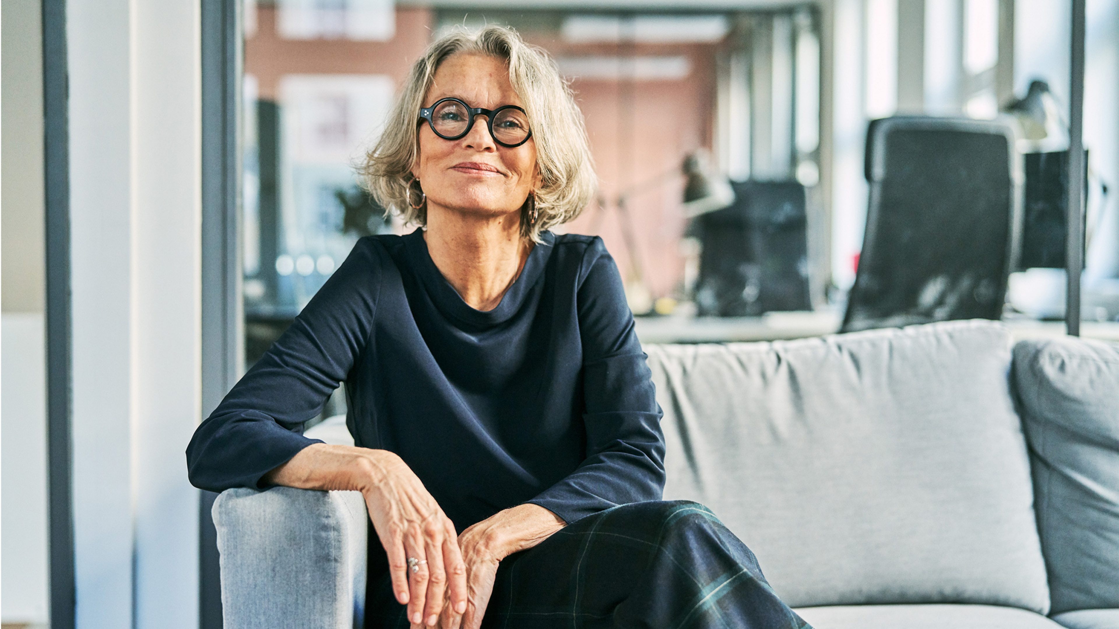 Une femme élégante d’une cinquantaine d’années, aux cheveux gris frisés et avec des lunettes rondes, est assise dans une posture décontractée sur un canapé gris dans un bureau moderne. Elle porte un haut bleu foncé et un pantalon à carreaux, elle sourit avec confiance et regarde directement la caméra. A l’arrière-plan, on peut voir des baies vitrées, du mobilier de bureau et des silhouettes floues de personnes au travail.