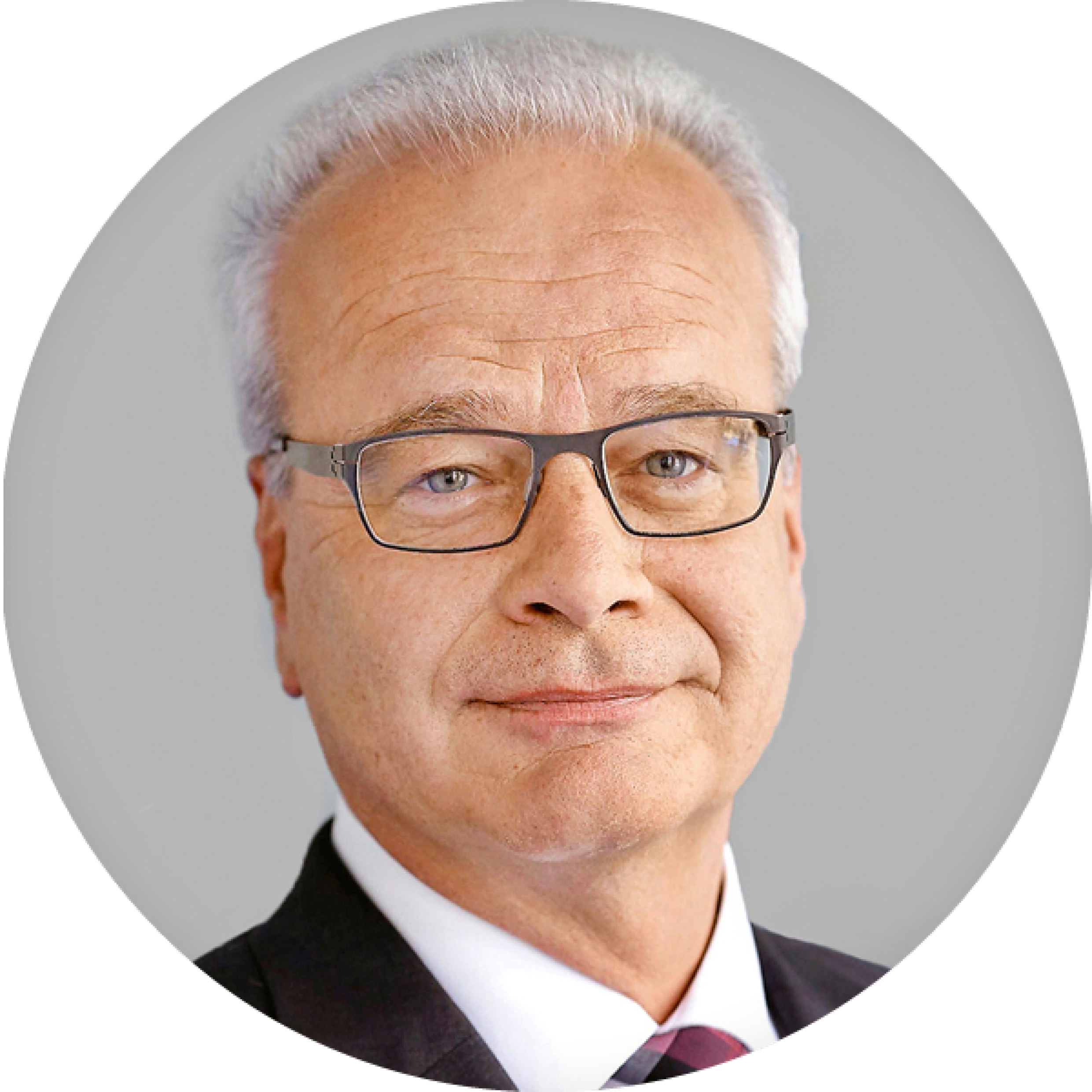 Un homme aux cheveux gris avec des yeux gris clair porte des lunettes et un costume. Il regarde la caméra en souriant.
