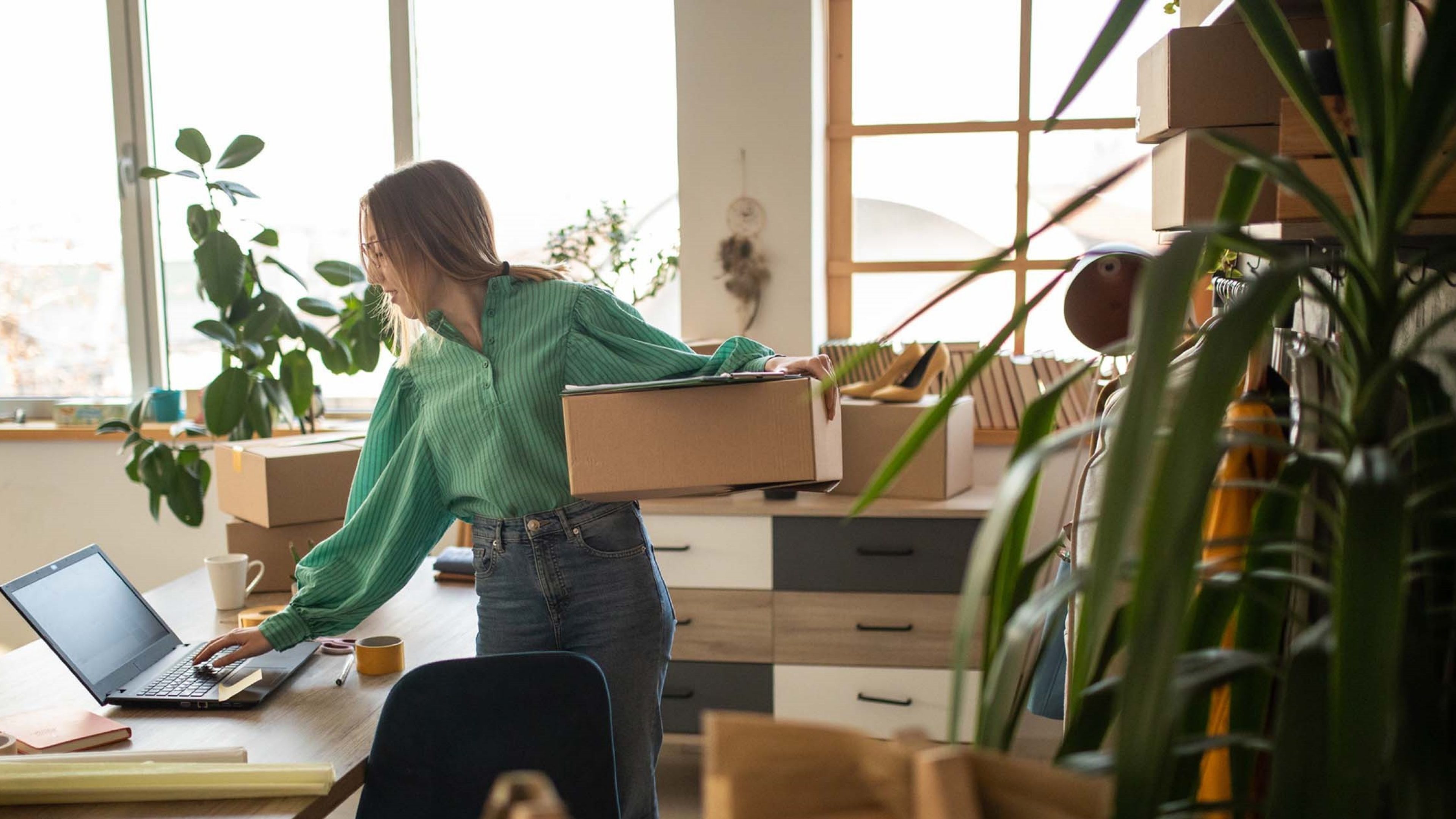 Une jeune femme blonde se tient debout dans un bureau lumineux et baigné de lumière. Elle tape d’une main sur le clavier d’un ordinateur portable posé sur une table et tient une boîte en carton dans l’autre main. D’autres boîtes en carton ainsi que plusieurs plantes vertes d’intérieur se trouvent autour d’elle. Elle porte un chemisier vert et un jean bleu.