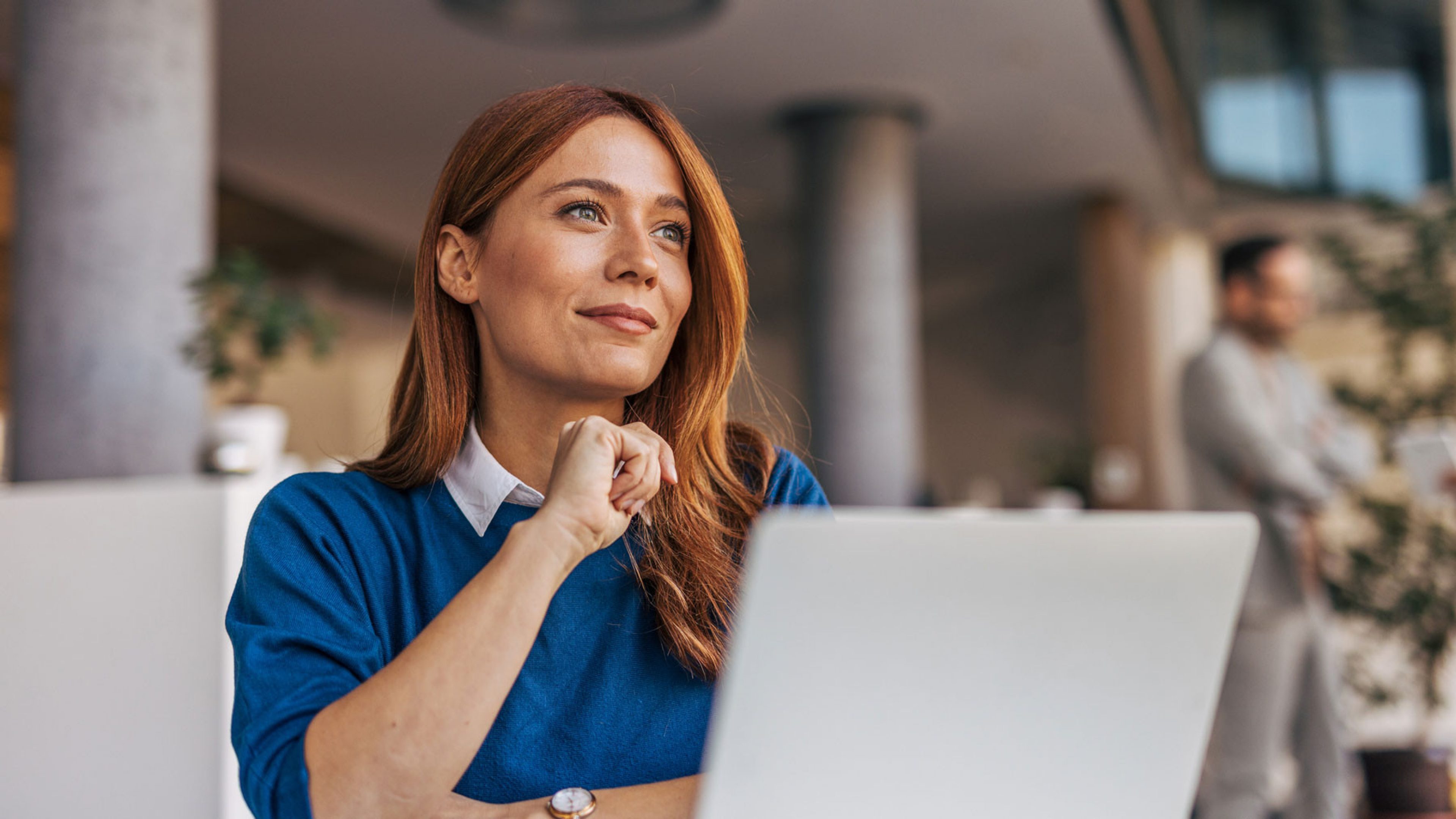 Une femme aux cheveux roux porte un sweat-shirt bleu et est assise devant un ordinateur portable, le regard au loin. En arrière-plan, on peut voir des meubles de bureau flous et une autre personne, un homme. L’ambiance est calme et professionnelle. 