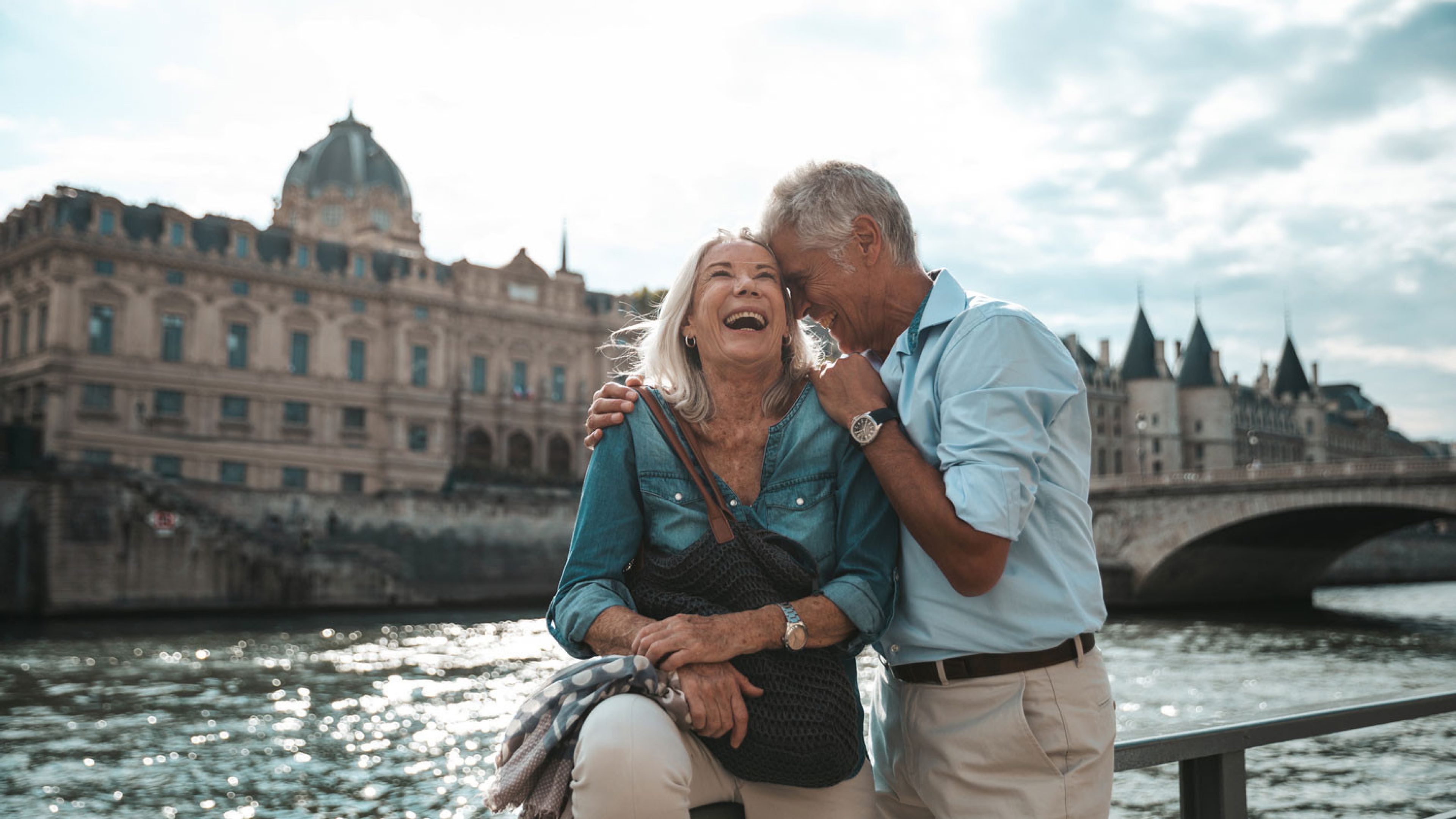 Un couple de personnes âgées se tient debout sur une balustrade en face d’une rivière, avec en arrière-plan un pont et un bâtiment historique. L’homme enlace la femme et lui murmure quelque chose à l’oreille. Tous deux portent des vêtements décontractés et rient de bon cœur.