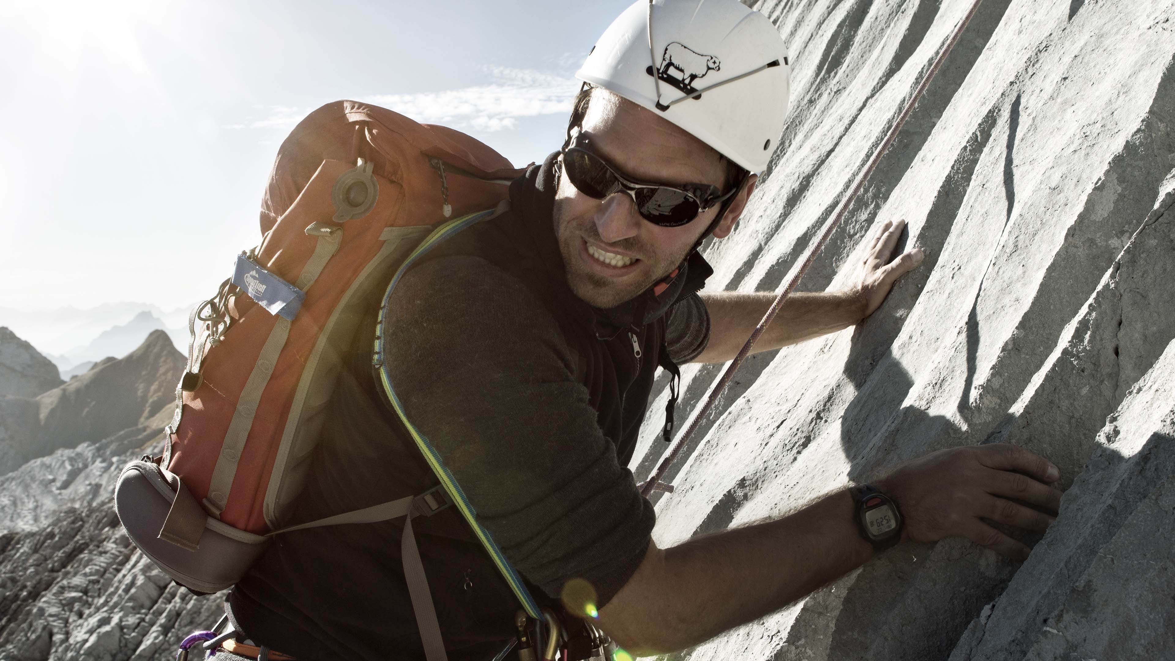 Personne escalade une paroi rocheuse avec casque et sac rouge, assurée par des cordes, montagnes et ciel derrière