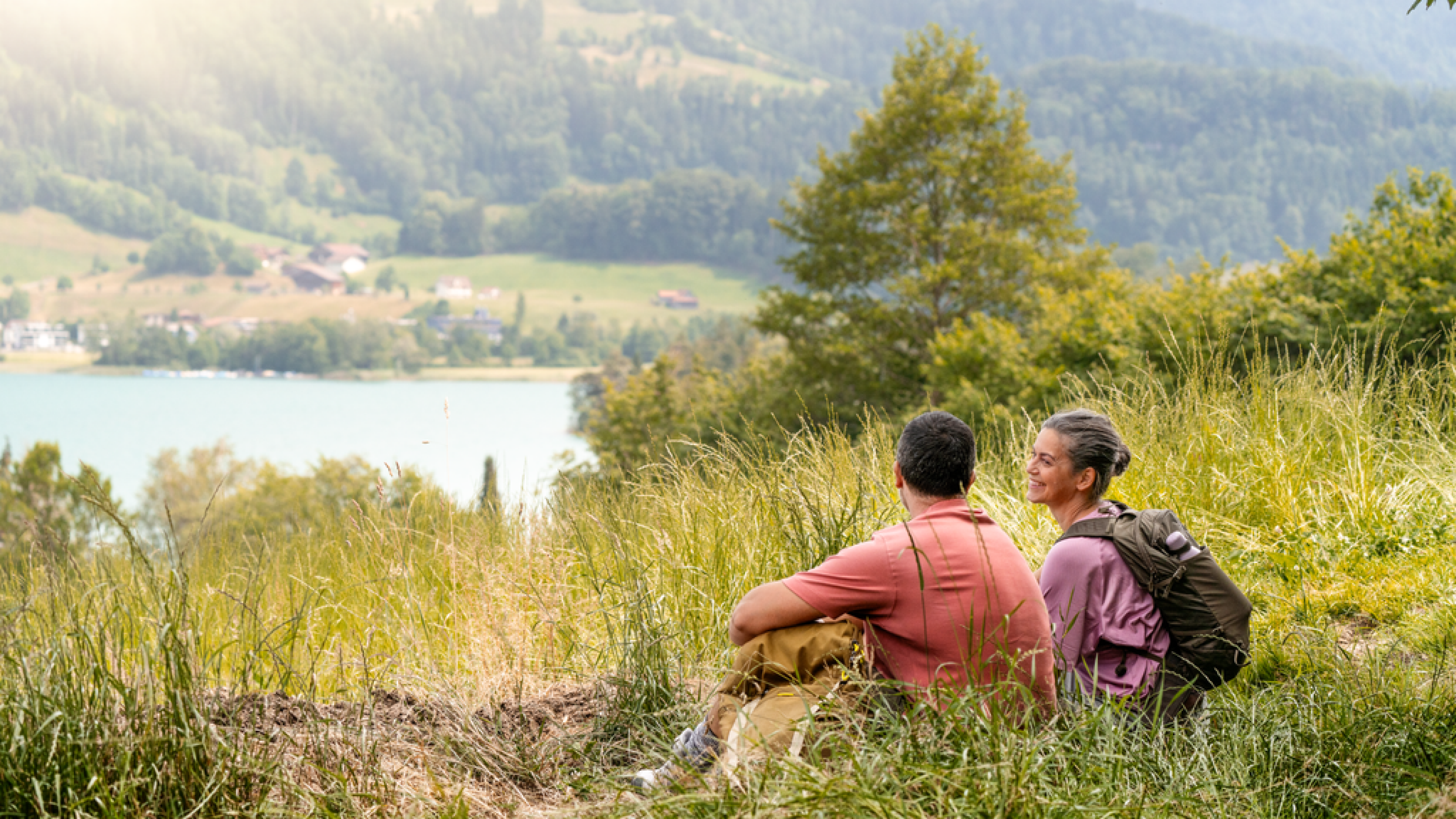Un couple de randonneurs s’accorde une pause dans l’herbe avec vue sur un lac.
