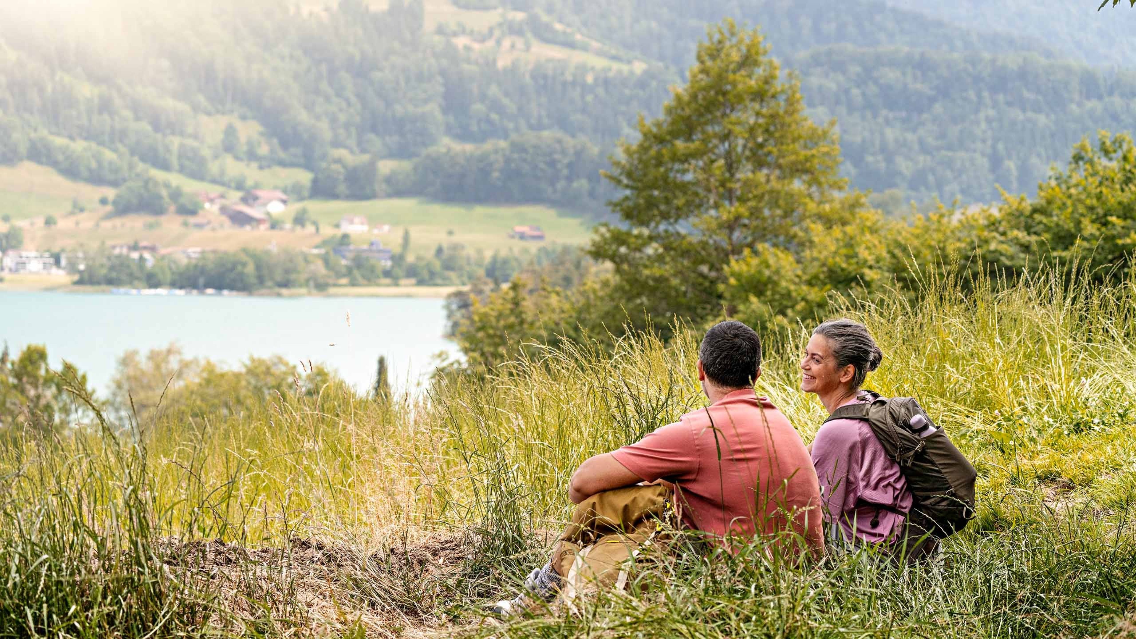 Un couple de randonneurs s’accorde une pause dans l’herbe avec vue sur un lac.