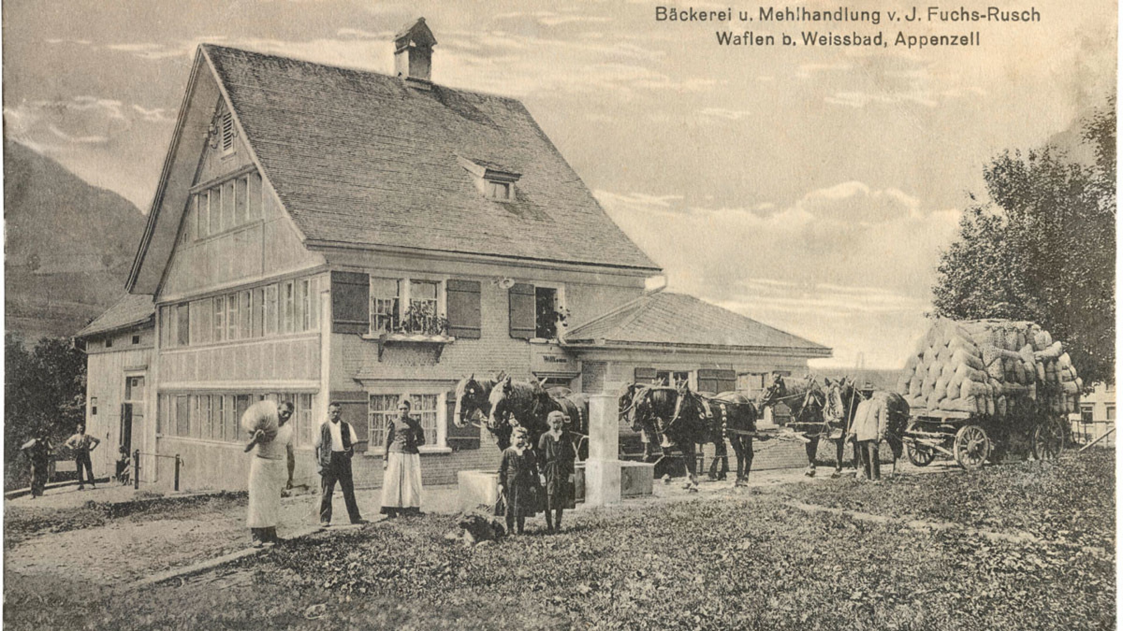 Photographie historique de Bischofberger AG. Plusieurs personnes en vêtements de l’époque se tiennent devant une maison de plusieurs étages avec un toit pentu. A droite de l’image, on peut voir une charrette avec des marchandises et tirée par des chevaux.