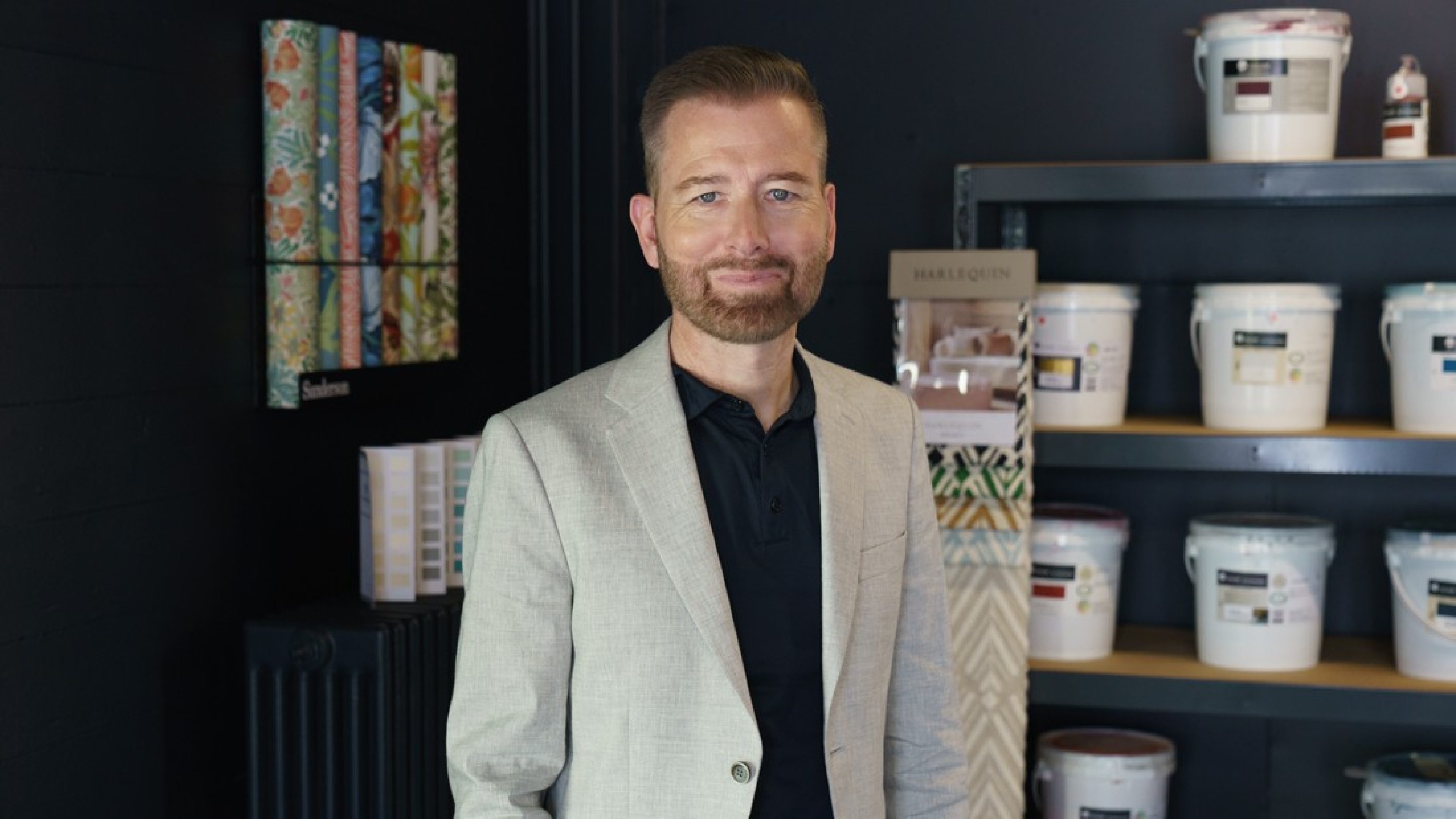 Derrière un homme en veste grise, des motifs de couleurs et de papier peint ornent les murs d’un showroom. Les motifs servent à la fois d’échantillons visuels et de décoration.