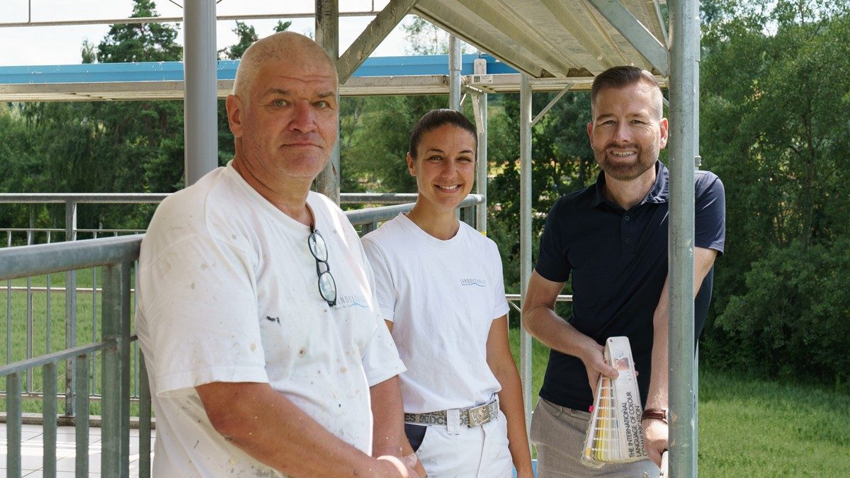 Un homme se tient à l’extérieur avec un nuancier de couleurs à la main et deux personnes sont à côté de lui sur un échafaudage. Ces personnes sont entièrement habillées en blanc..