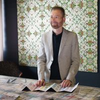 Un homme vêtu d’une veste grise a déplié un nuancier de couleurs sur une table. En arrière-plan, un papier peint attire l’attention: des feuilles et des papillons y sont répartis de manière symétrique comme dans un kaléidoscope.