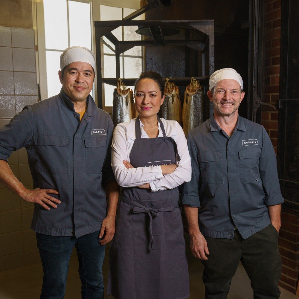 Trois personnes en vêtements de travail se trouvent devant les filets de saumon suspendus et sourient à la caméra. Deux hommes et une femme au milieu. Les hommes portent des chemises bleu foncé et des toques blanches; la femme porte un tablier et un chemisier blanc et se tient les bras croisés.